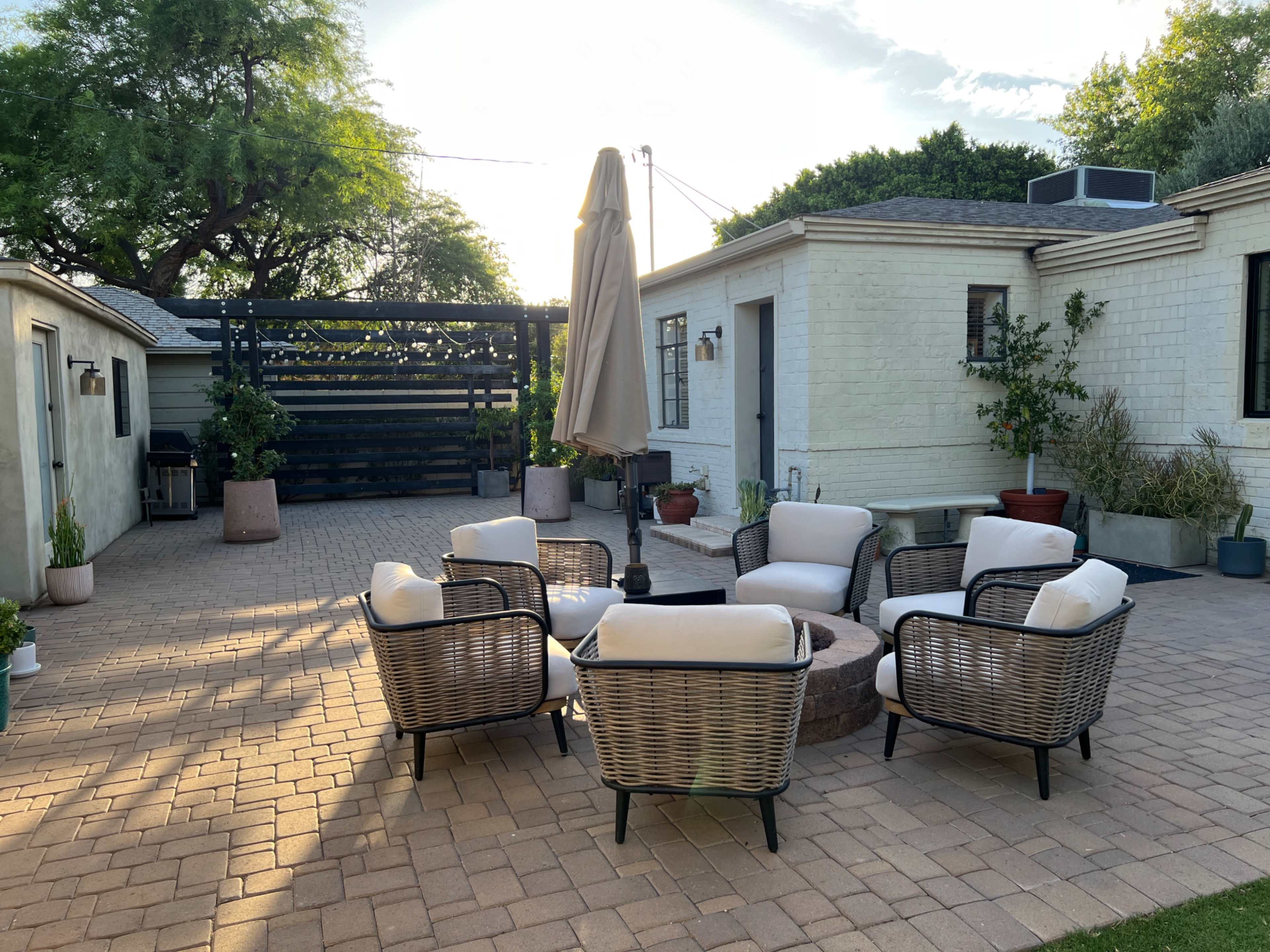 A patio area features a circular seating arrangement with four wicker chairs and a table surrounding a fire pit, with a sun umbrella and potted plants in the background.