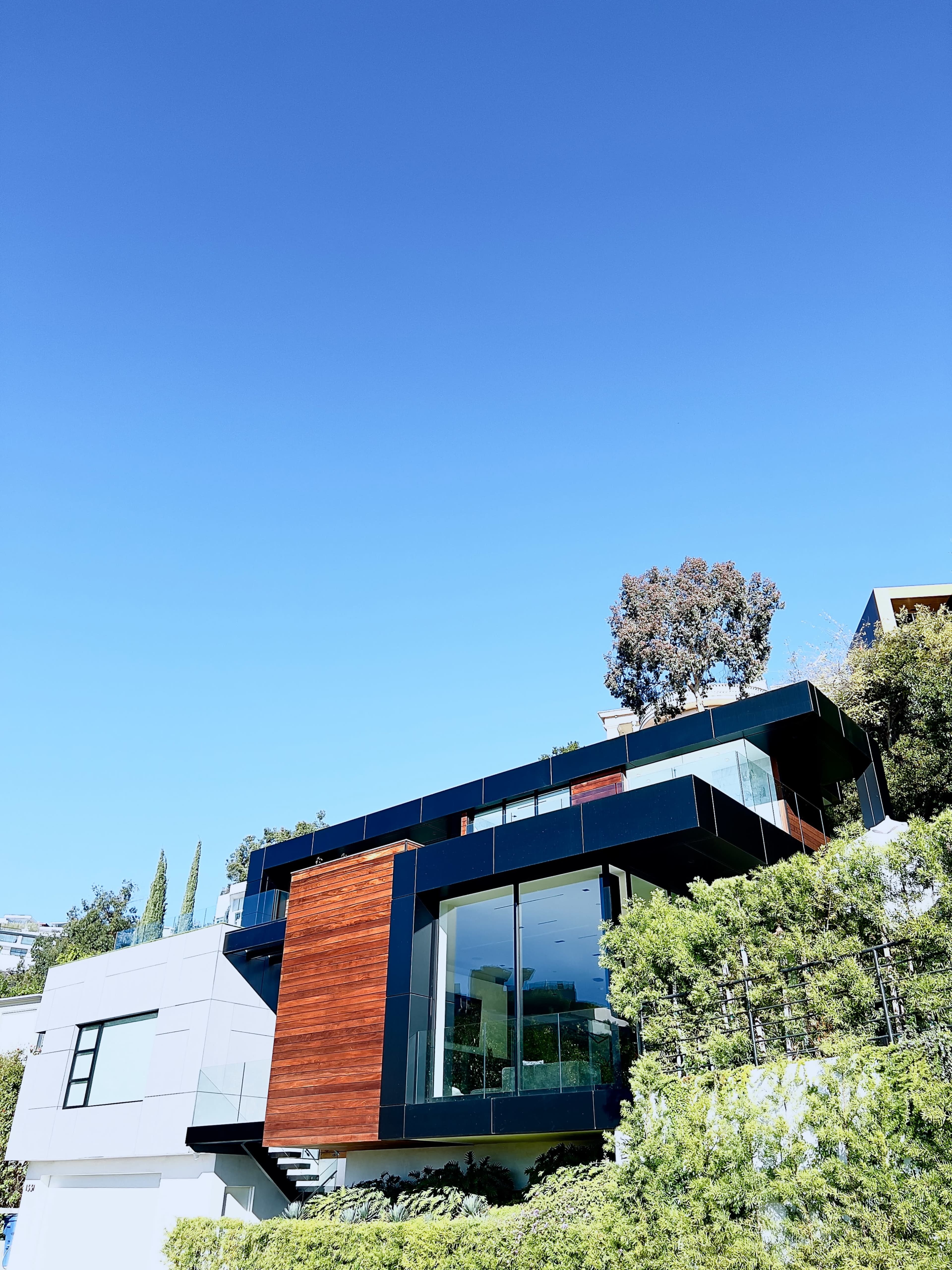 A modern house features large glass windows, wooden accents, and is surrounded by greenery under a clear blue sky.