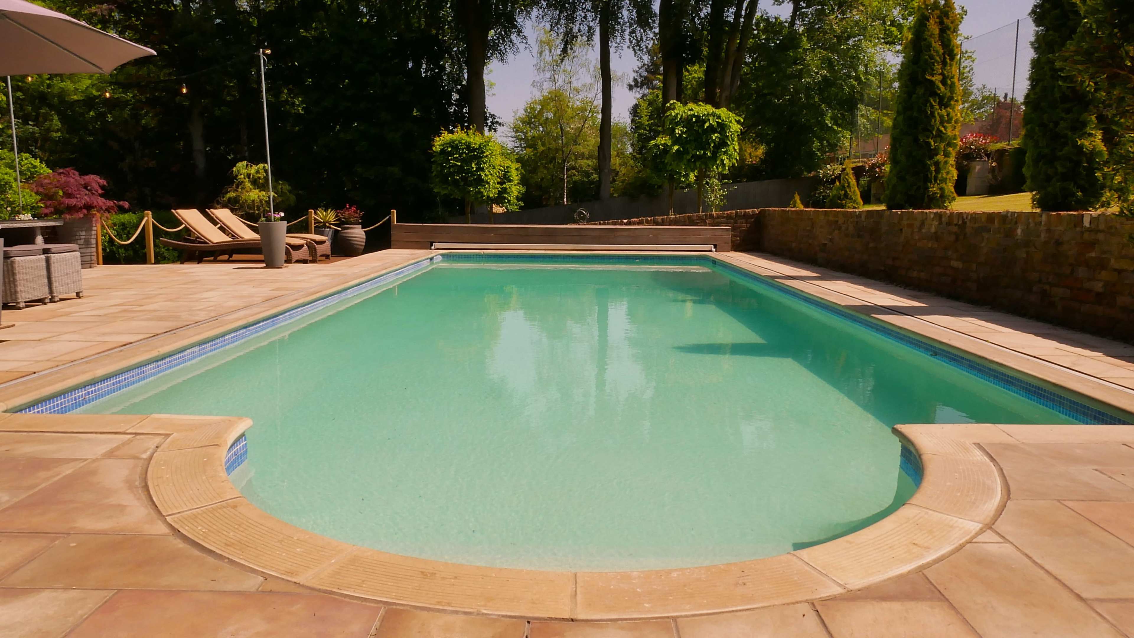 A clear swimming pool is surrounded by a stone patio and landscaped greenery.