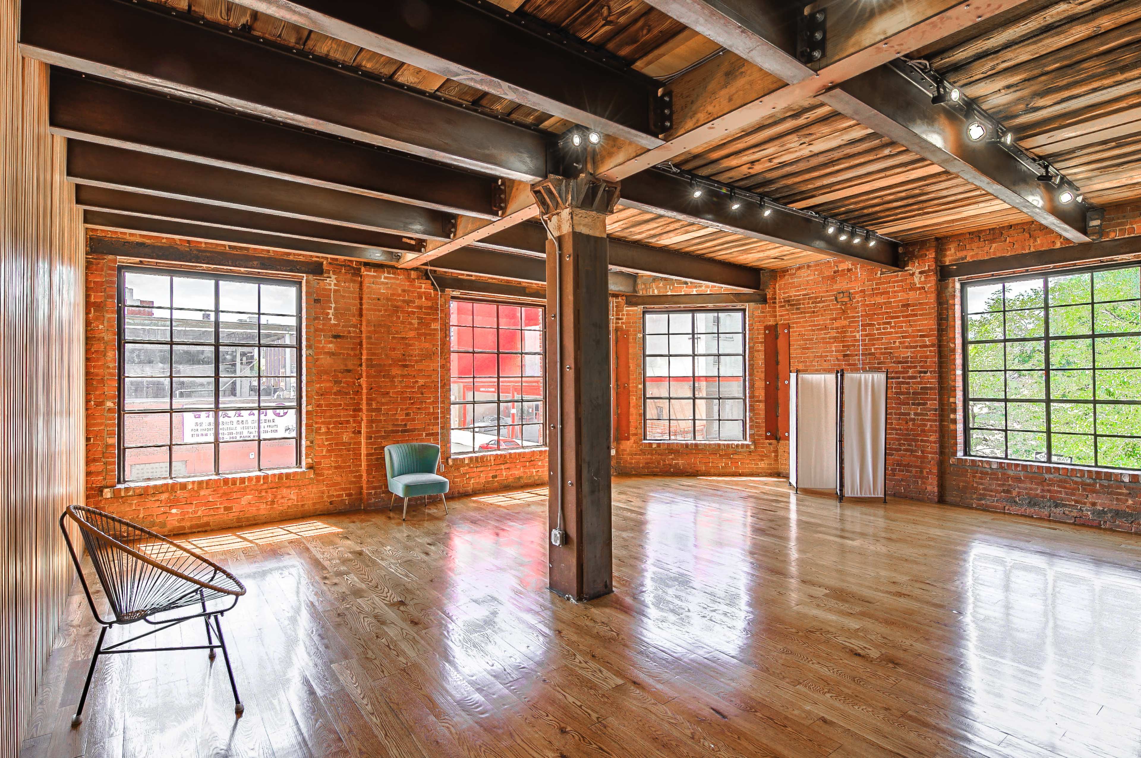The image shows a spacious room with exposed brick walls, wooden beams, and large windows allowing natural light to enter, alongside a green chair and a folding screen in the corner.
