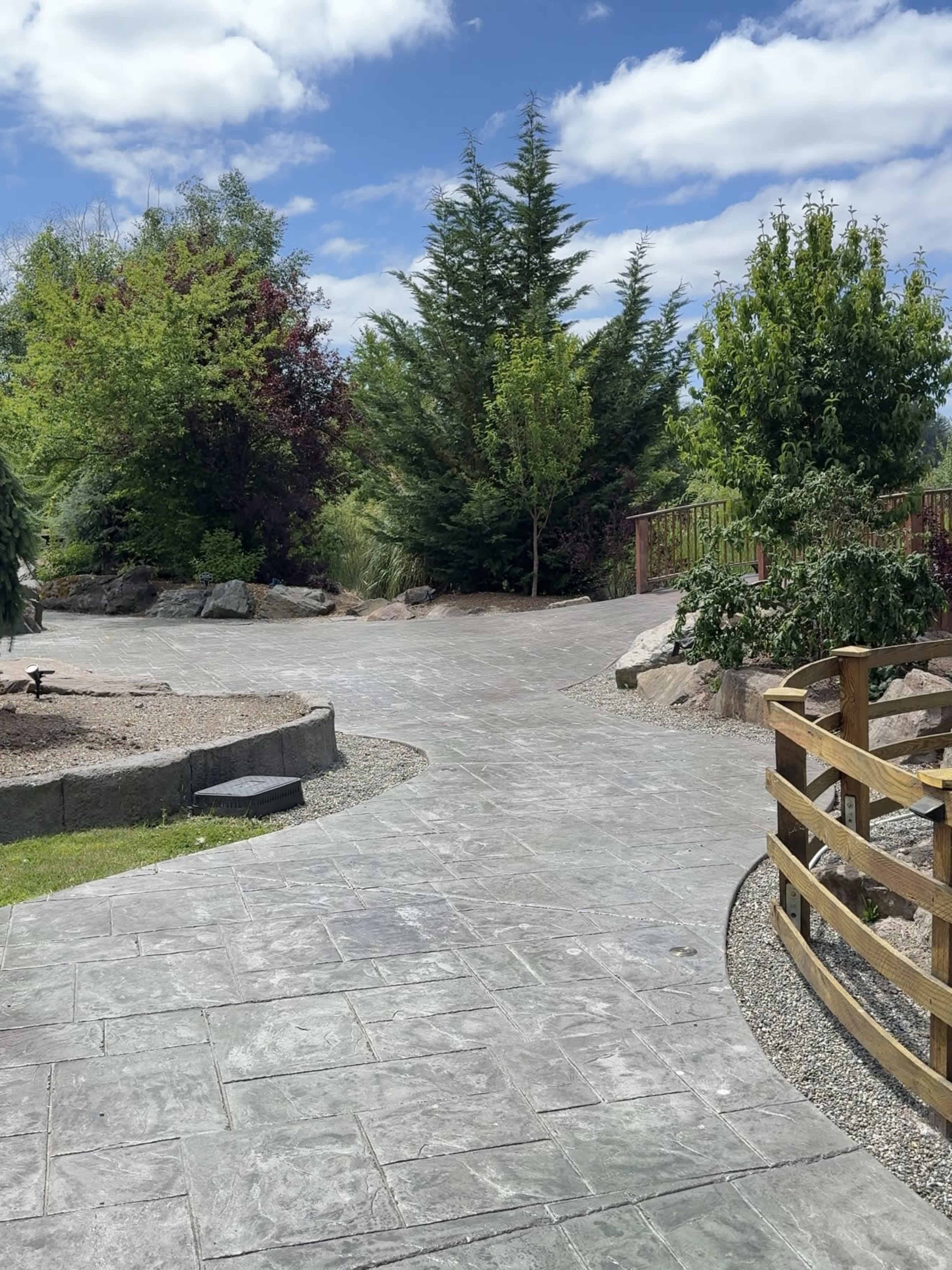 The image shows a landscaped pathway winding through greenery, featuring trees and a wooden fence under a partly cloudy sky.