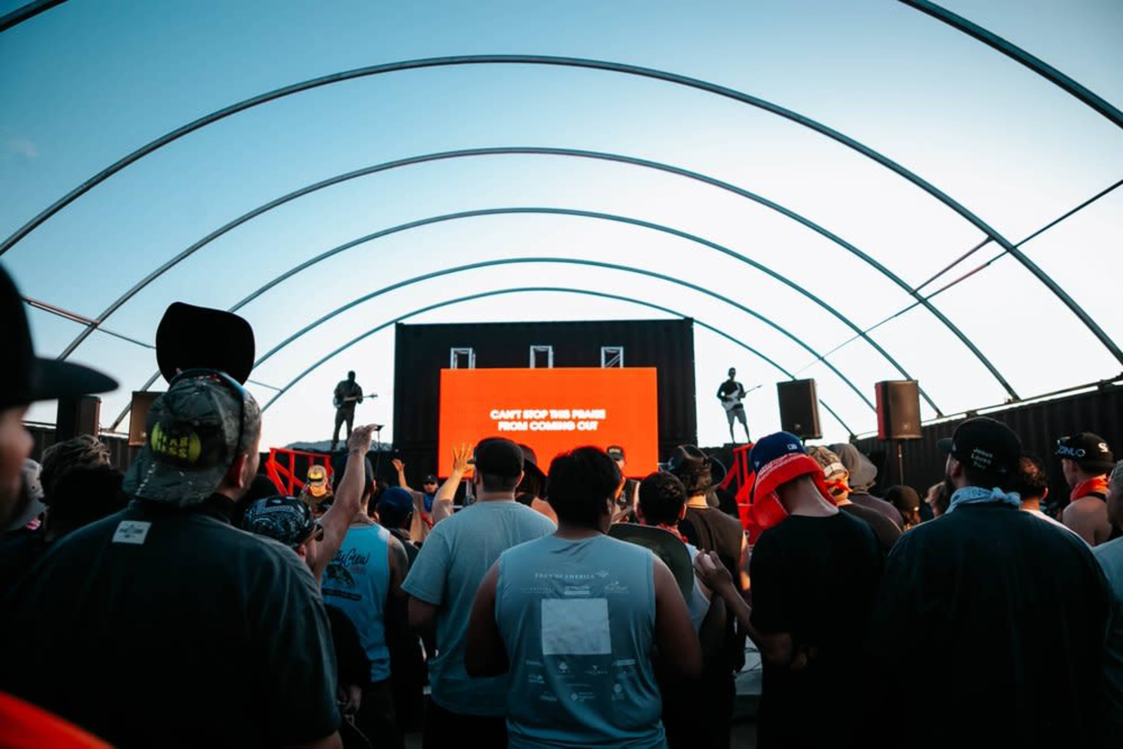 A crowd of people enjoys a live performance under a large, arching canopy with a brightly lit screen displaying a message.