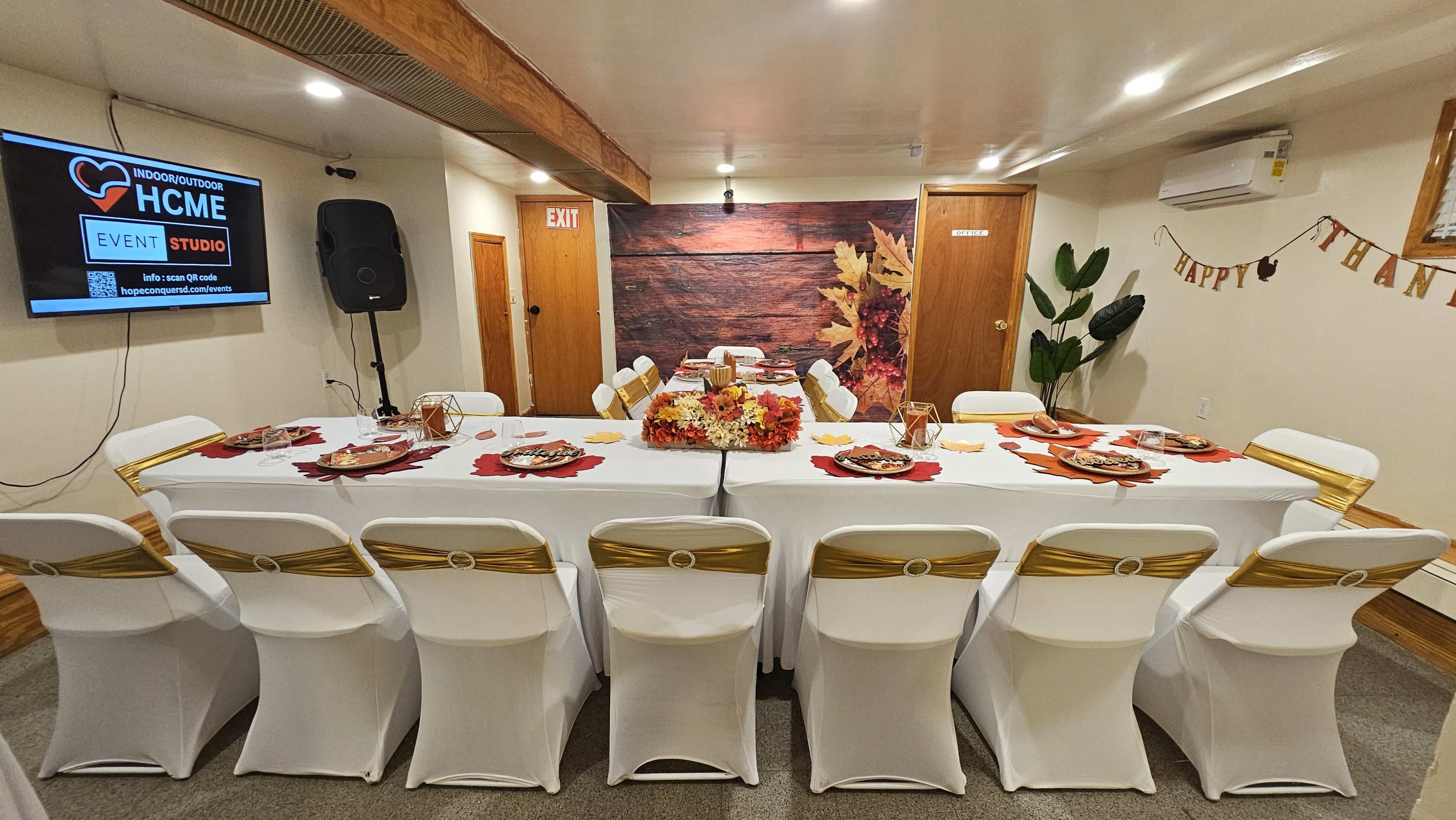 A long table is set for a festive meal in a decorated event space, with autumn-themed centerpieces and chairs arranged around it.