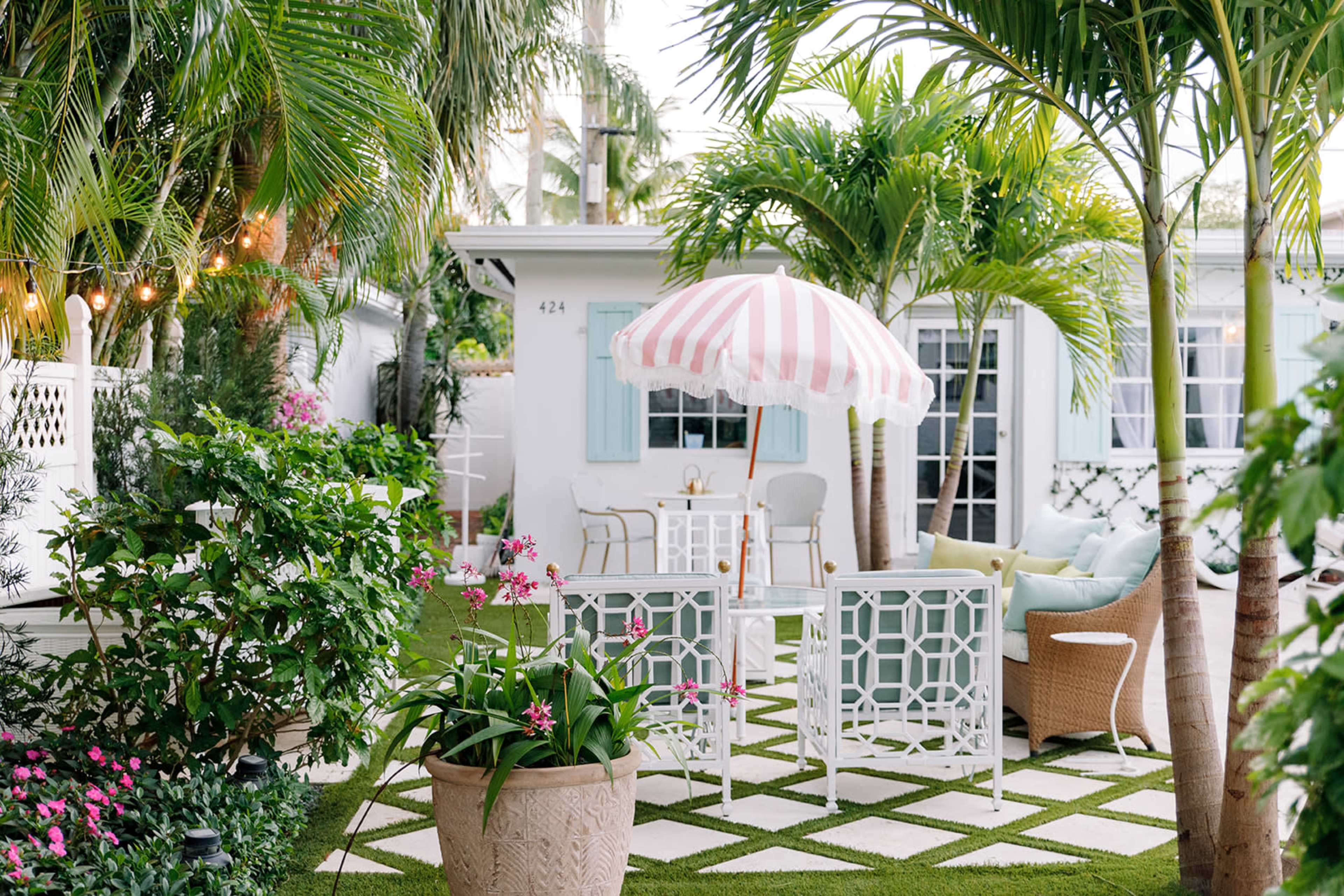 The image shows a garden area with white furniture, a pink and white striped umbrella, and various tropical plants surrounding a small table and chairs.