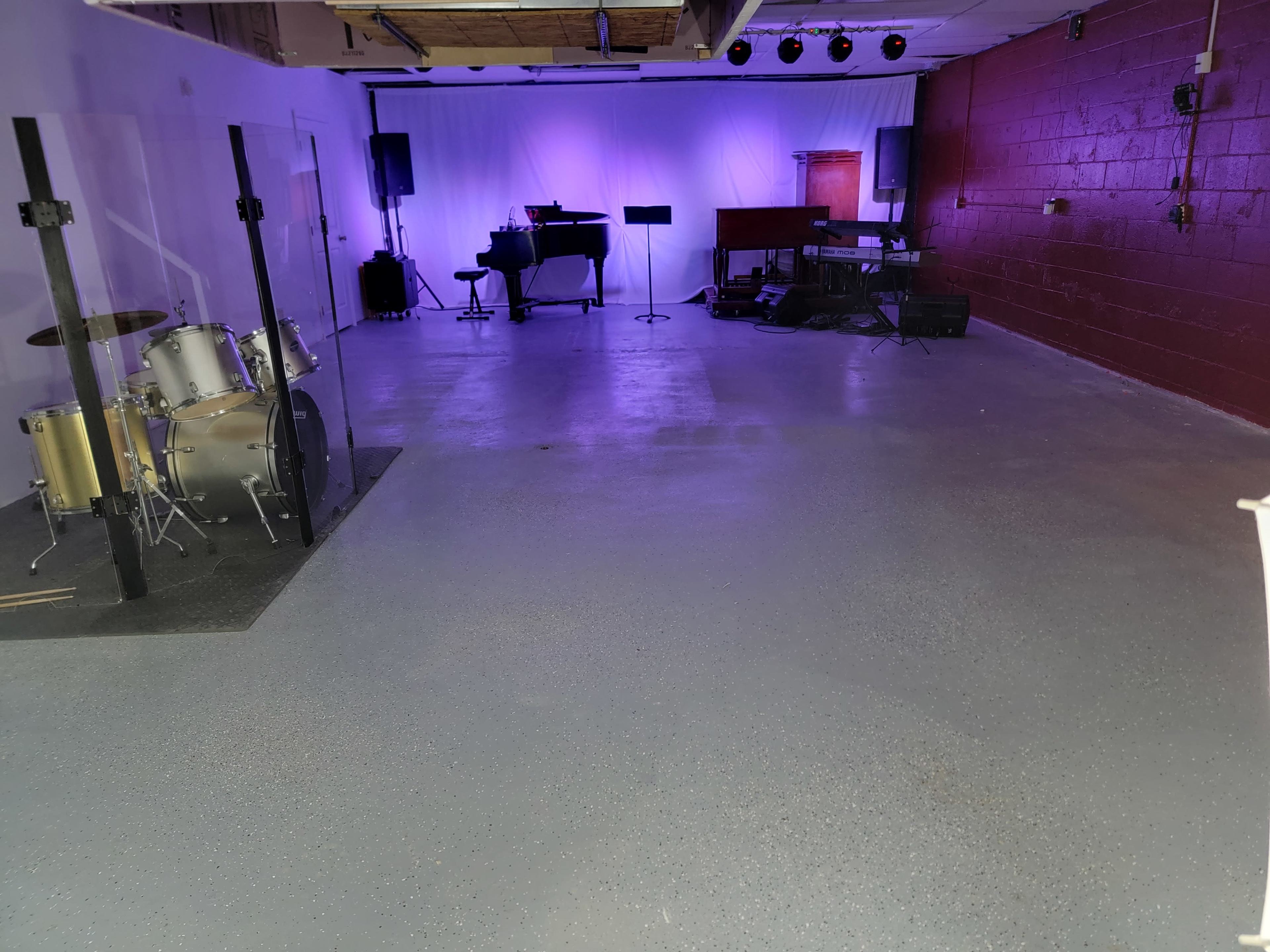 The image shows an empty room with set up musical instruments, including a drum set and a piano, with purple lighting in the background.
