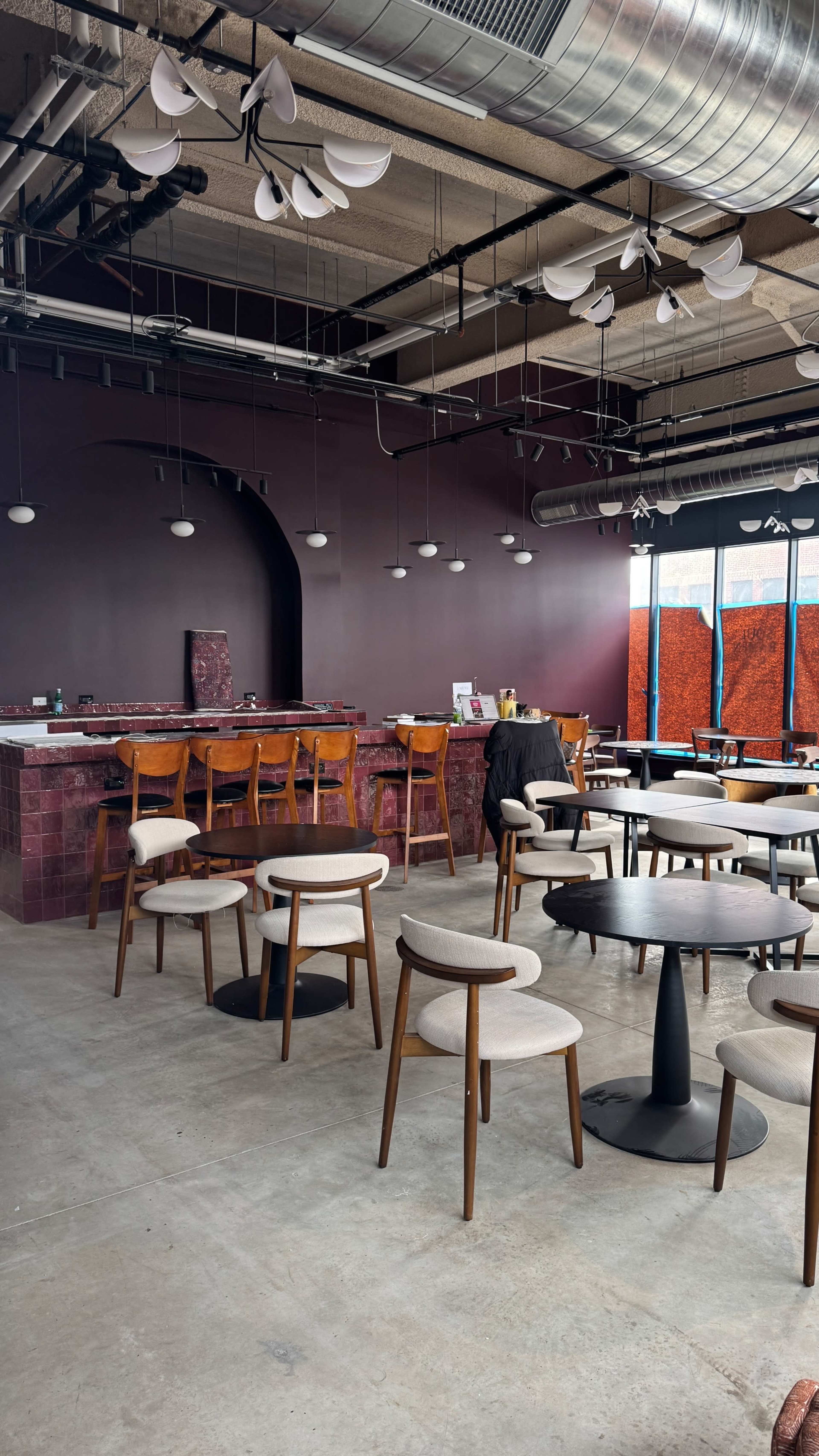 The image shows a modern café interior with wooden tables and chairs, a bar counter against a dark purple wall, and large windows that let in natural light.