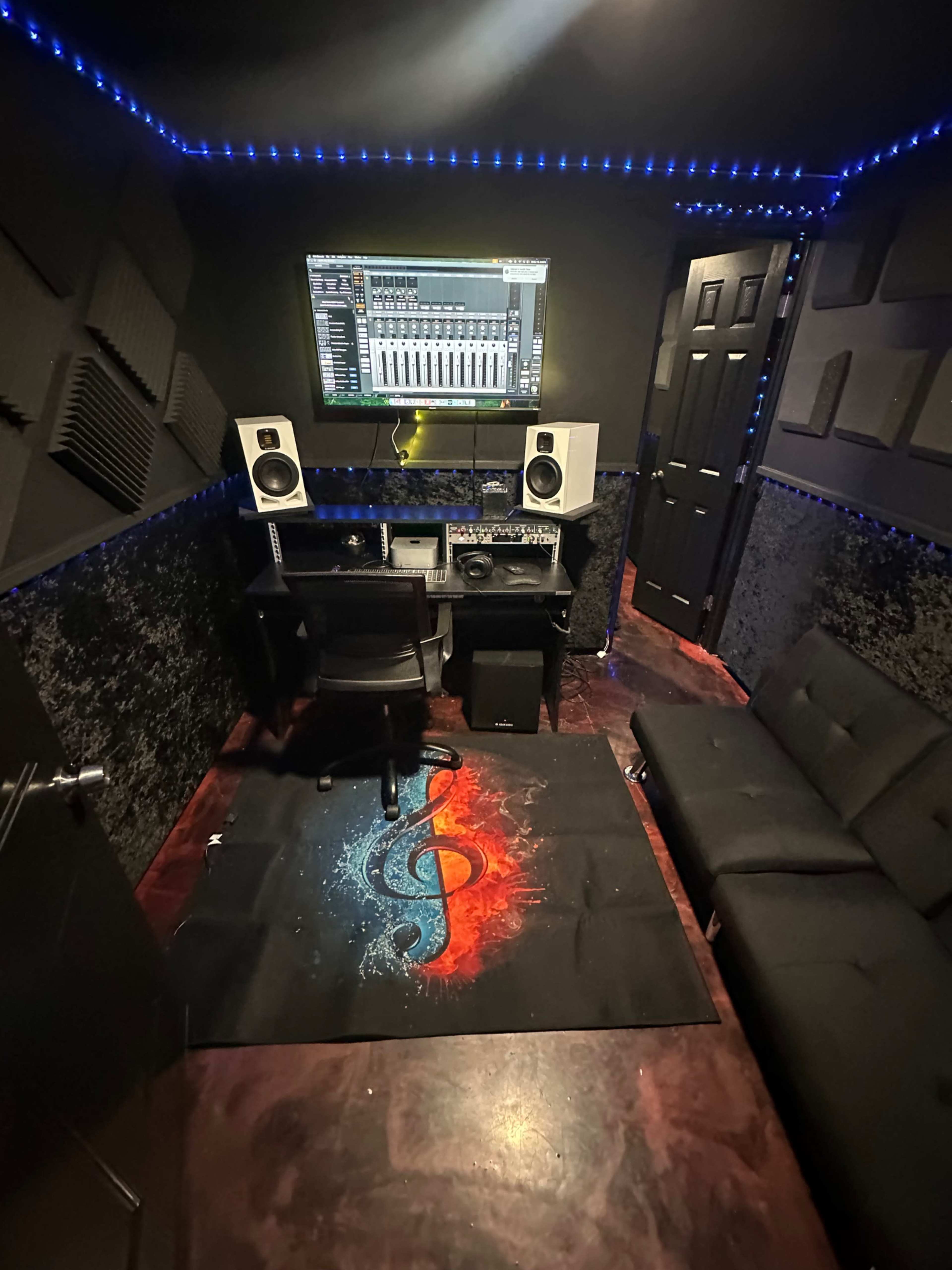 The image shows a small music production studio with a computer setup, speakers, and a colorful rug featuring a treble clef design.