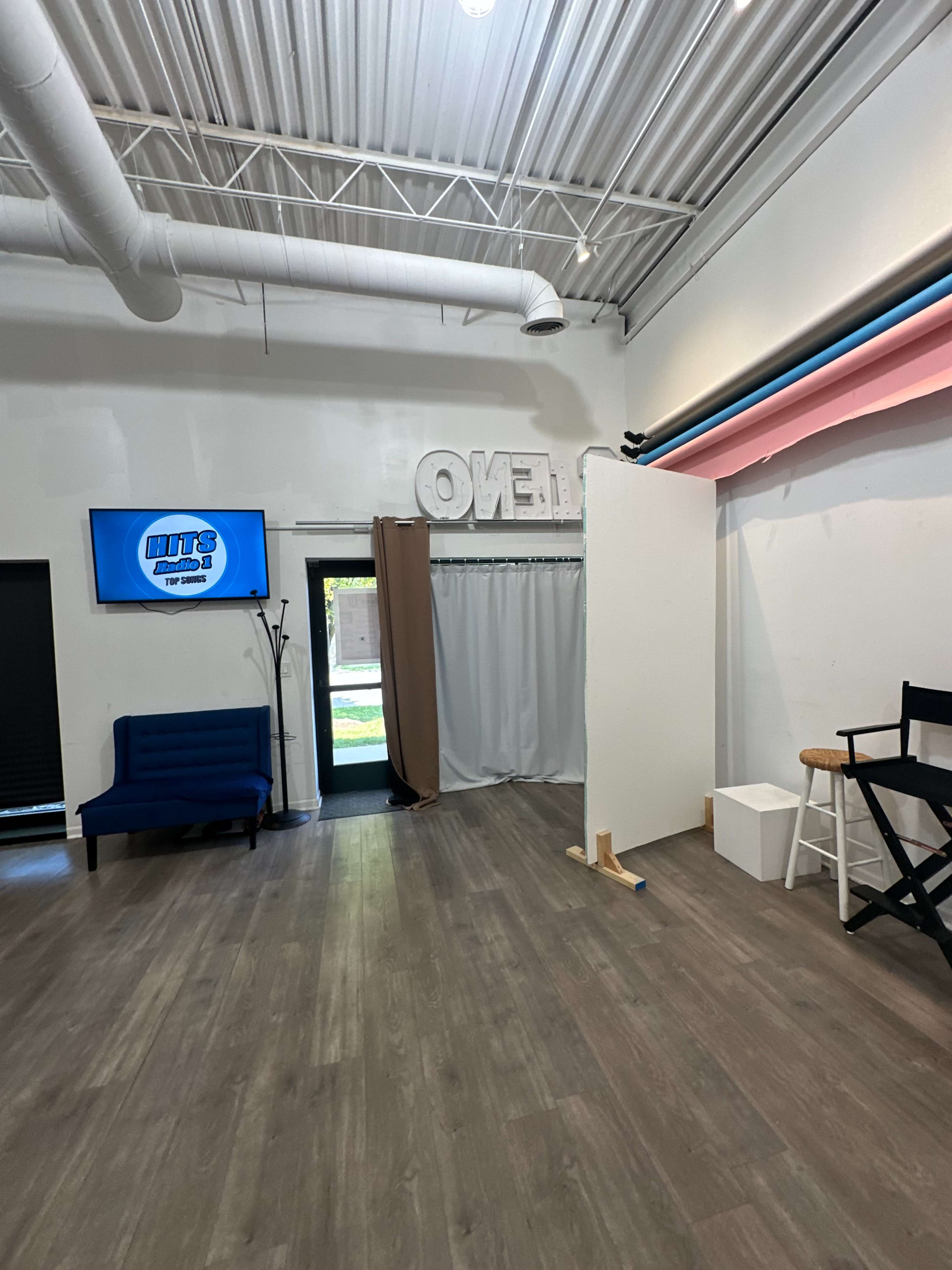 The image shows a bright, spacious room with a blue sofa, a television screen on the wall, and various setups for photography or filming.