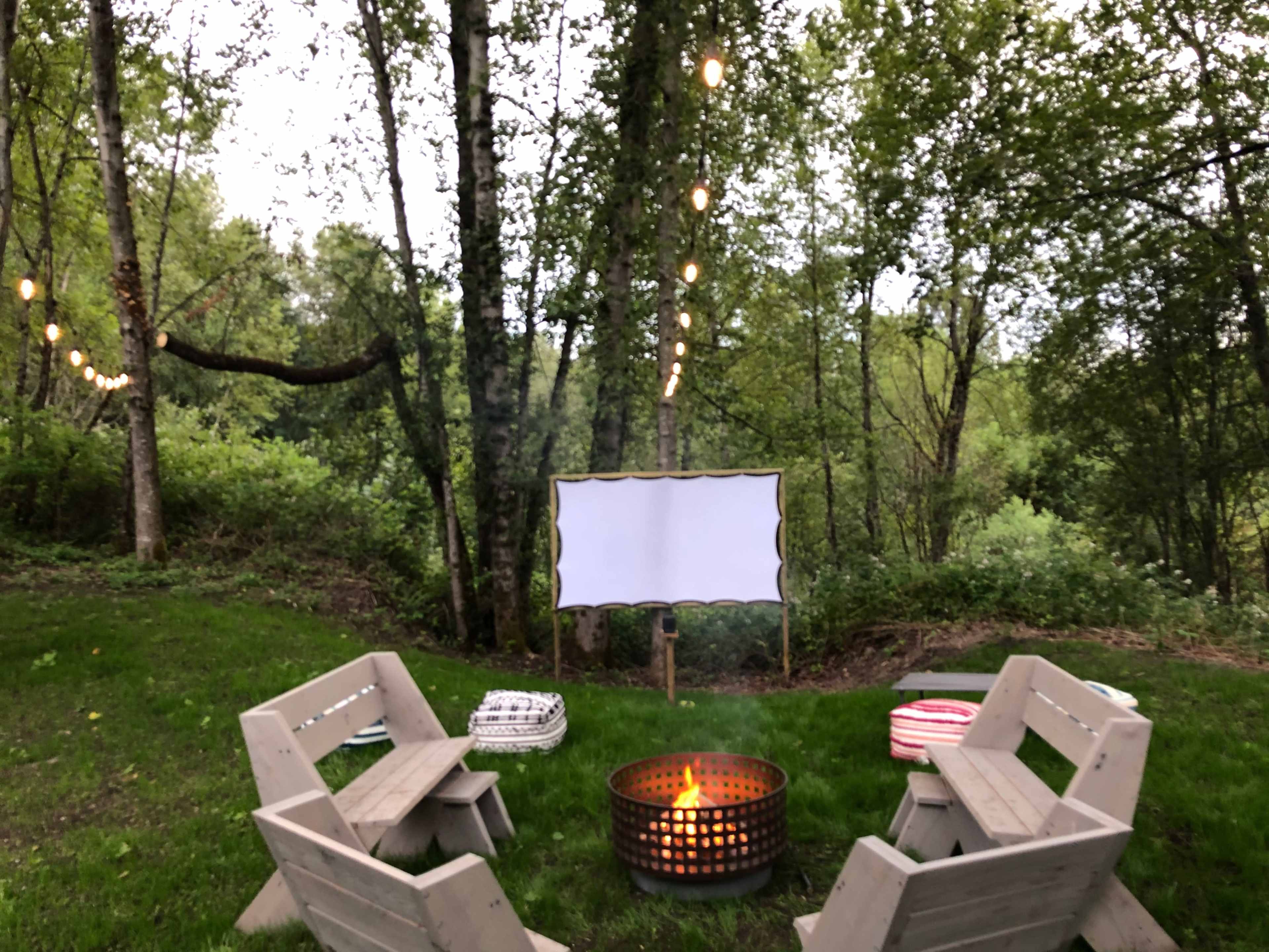The image shows a cozy outdoor setup featuring a circular fire pit surrounded by wooden seating, with string lights overhead and a blank screen positioned among trees in a green landscape.