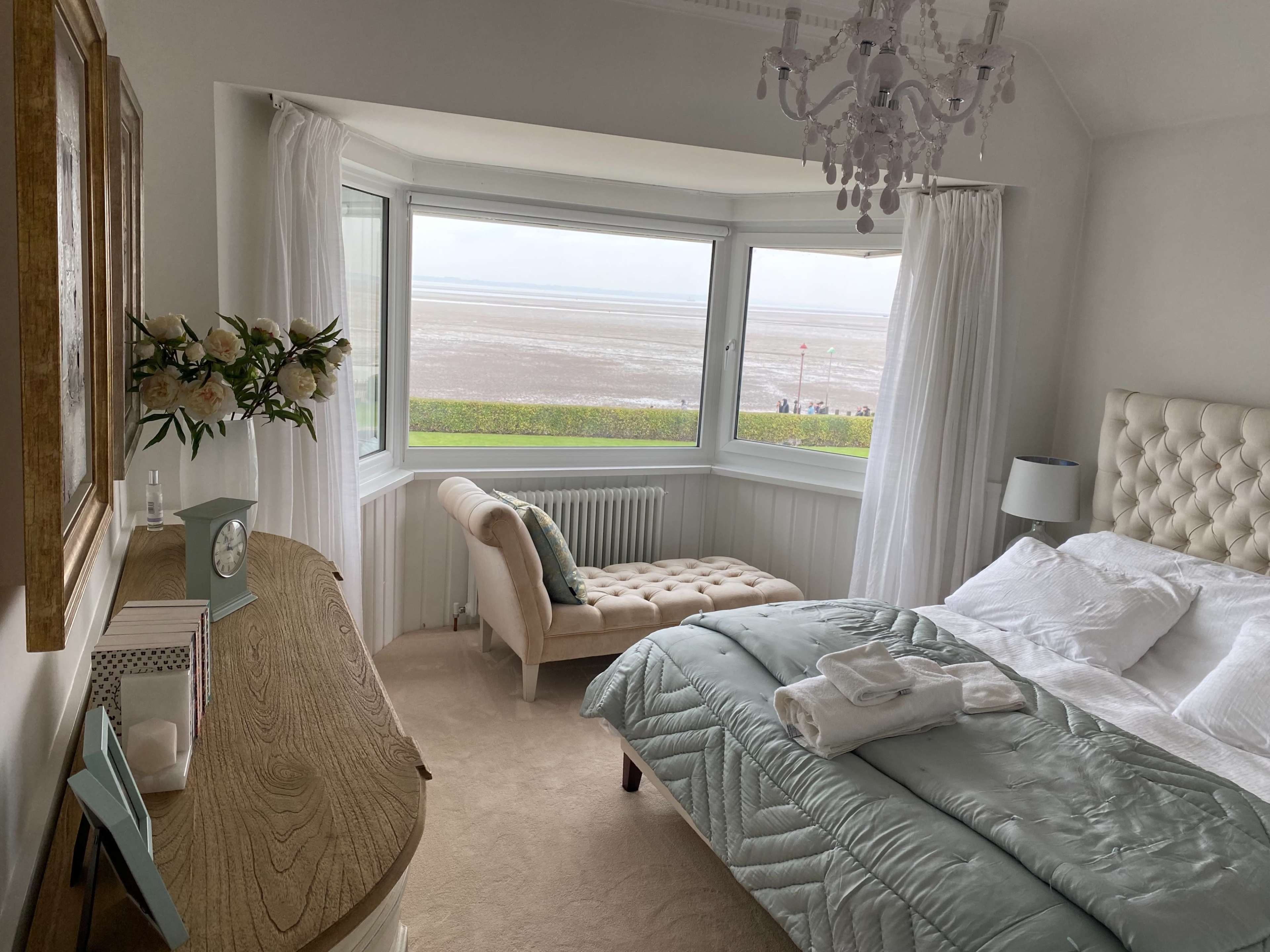 A well-decorated bedroom with a large window overlooking a beach, featuring a bed, a chaise lounge, and a wooden dresser.
