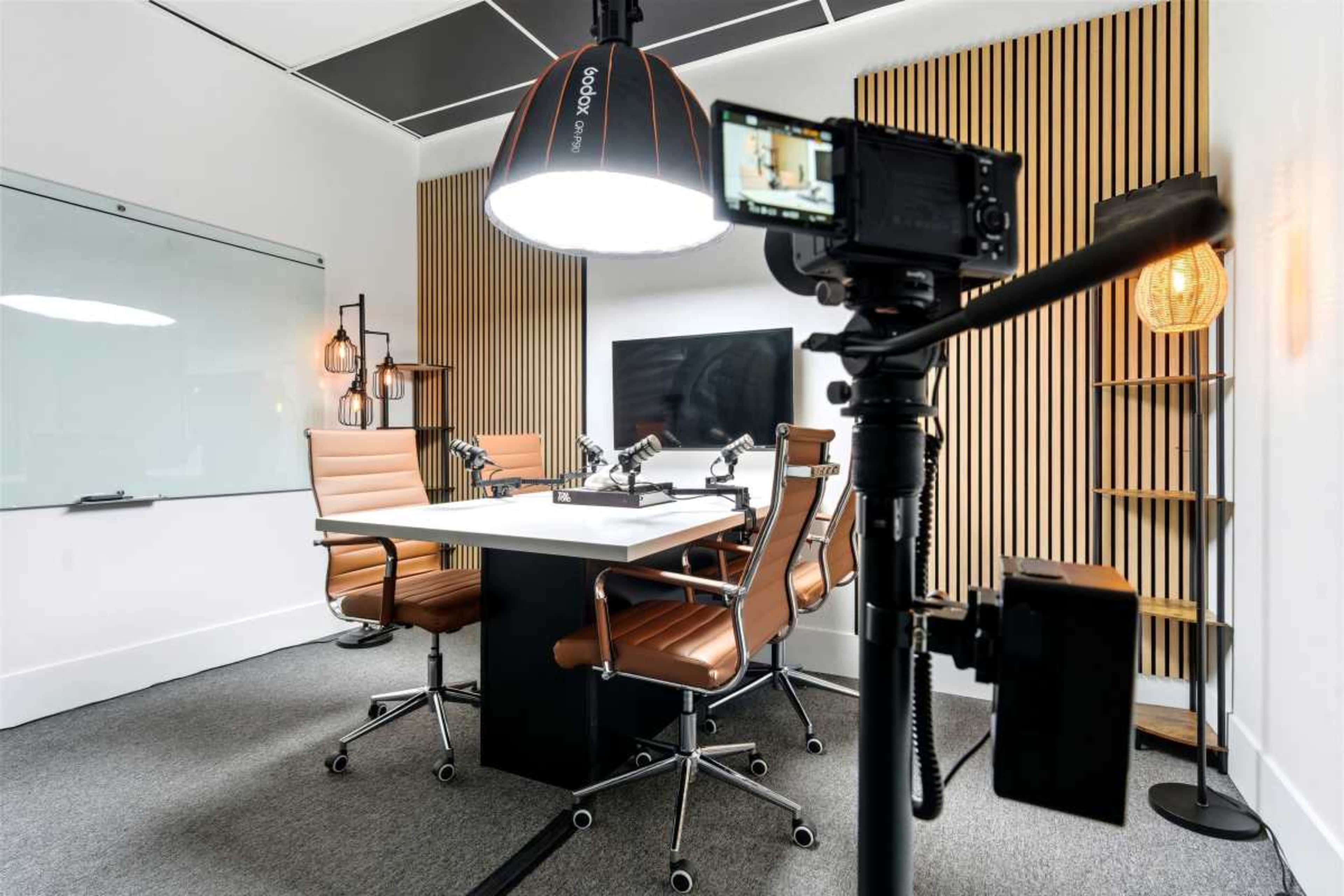 A modern conference room is equipped with a large central table, four brown leather chairs, and a camera mounted on a tripod under a large overhead light.