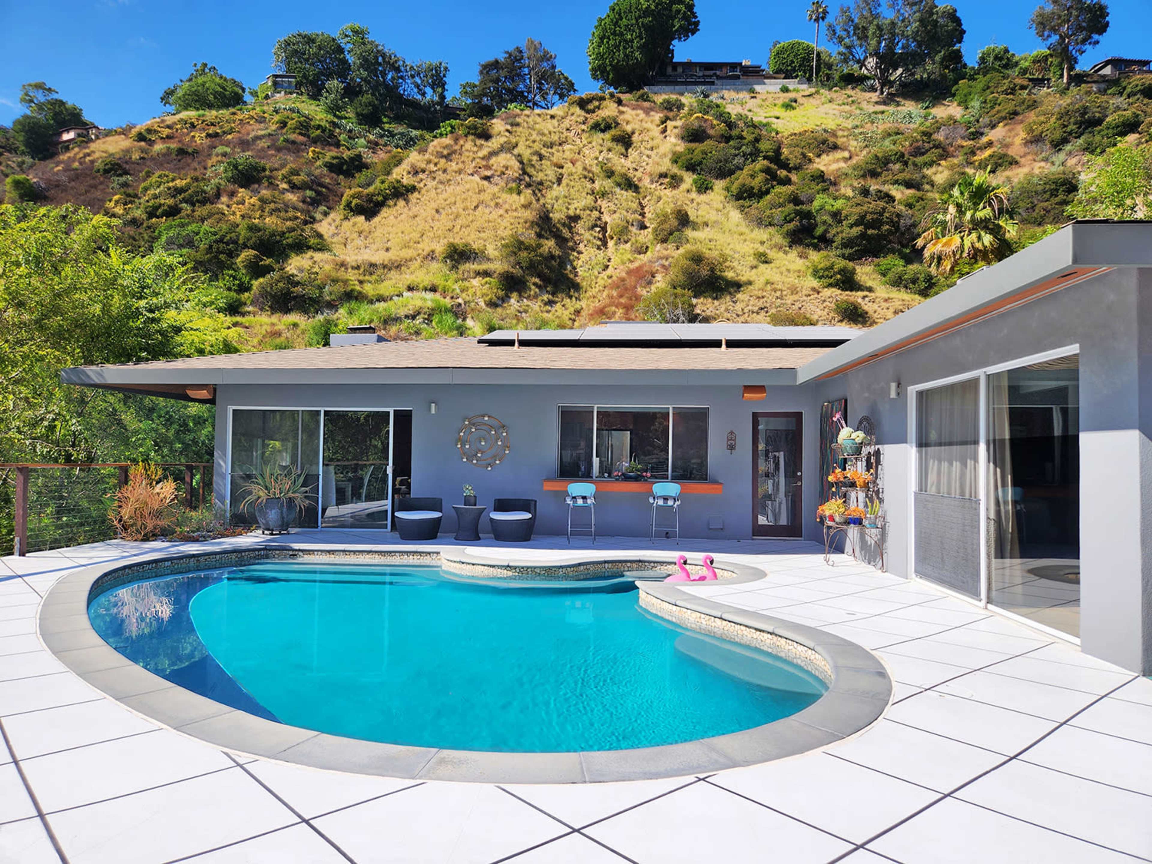 A modern house with a swimming pool, situated on a hillside with green vegetation in the background.