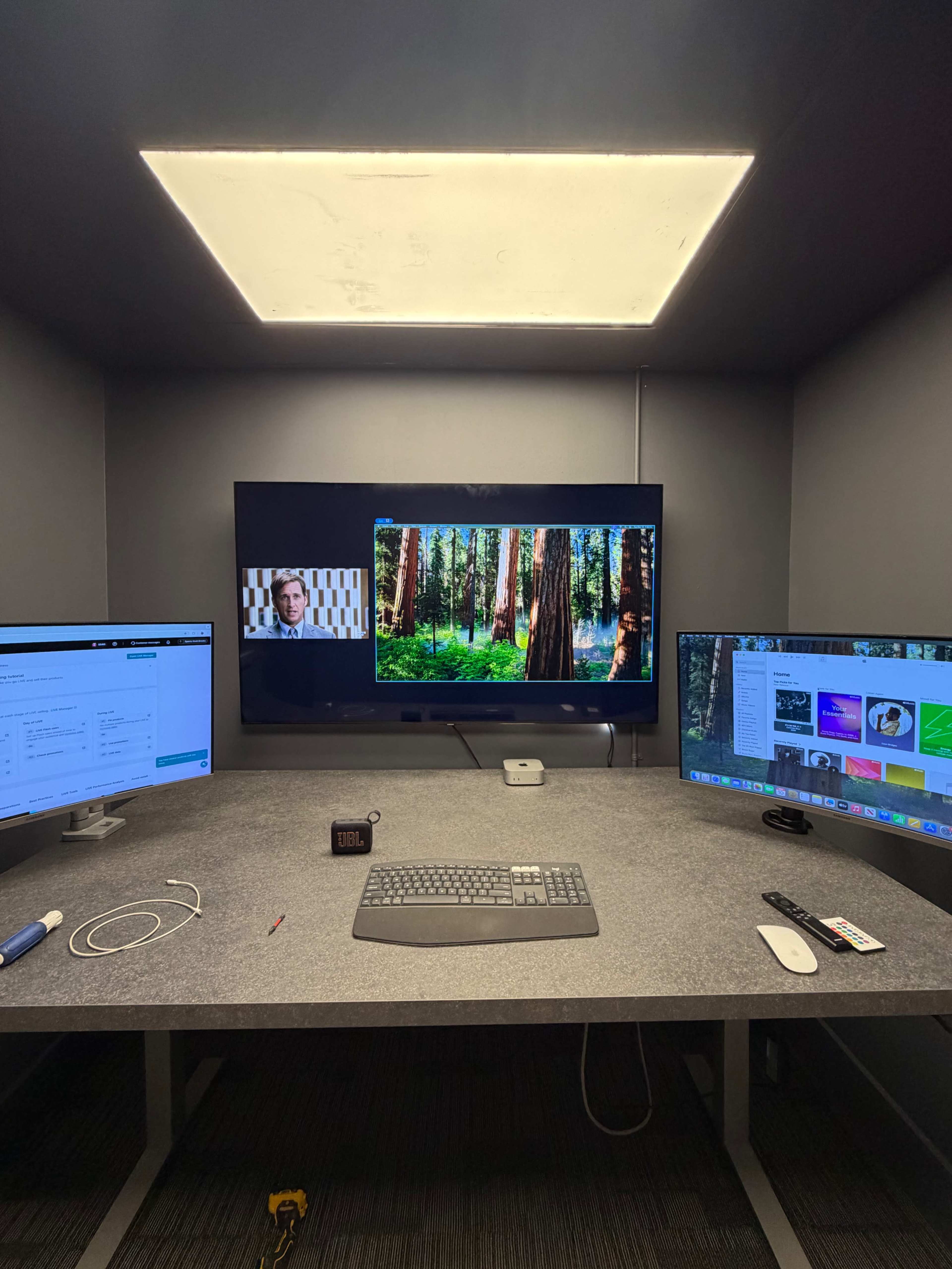 The image shows a modern workspace with two monitors on either side of a central screen displaying a forest scene, all set against a gray wall.