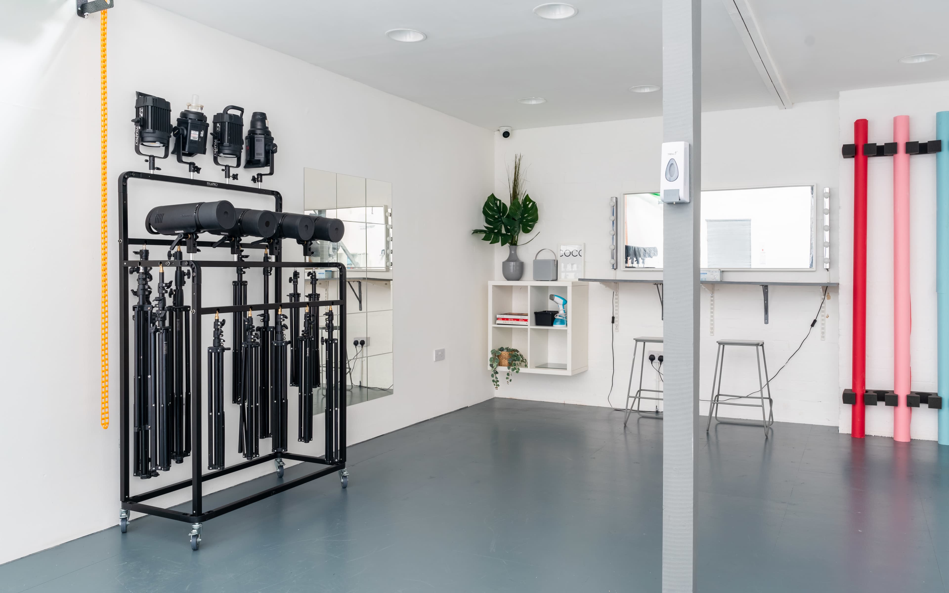 A minimalist studio space featuring lighting equipment on a rack, mirrors, a plant, and shelves with various items against a clean, white wall.