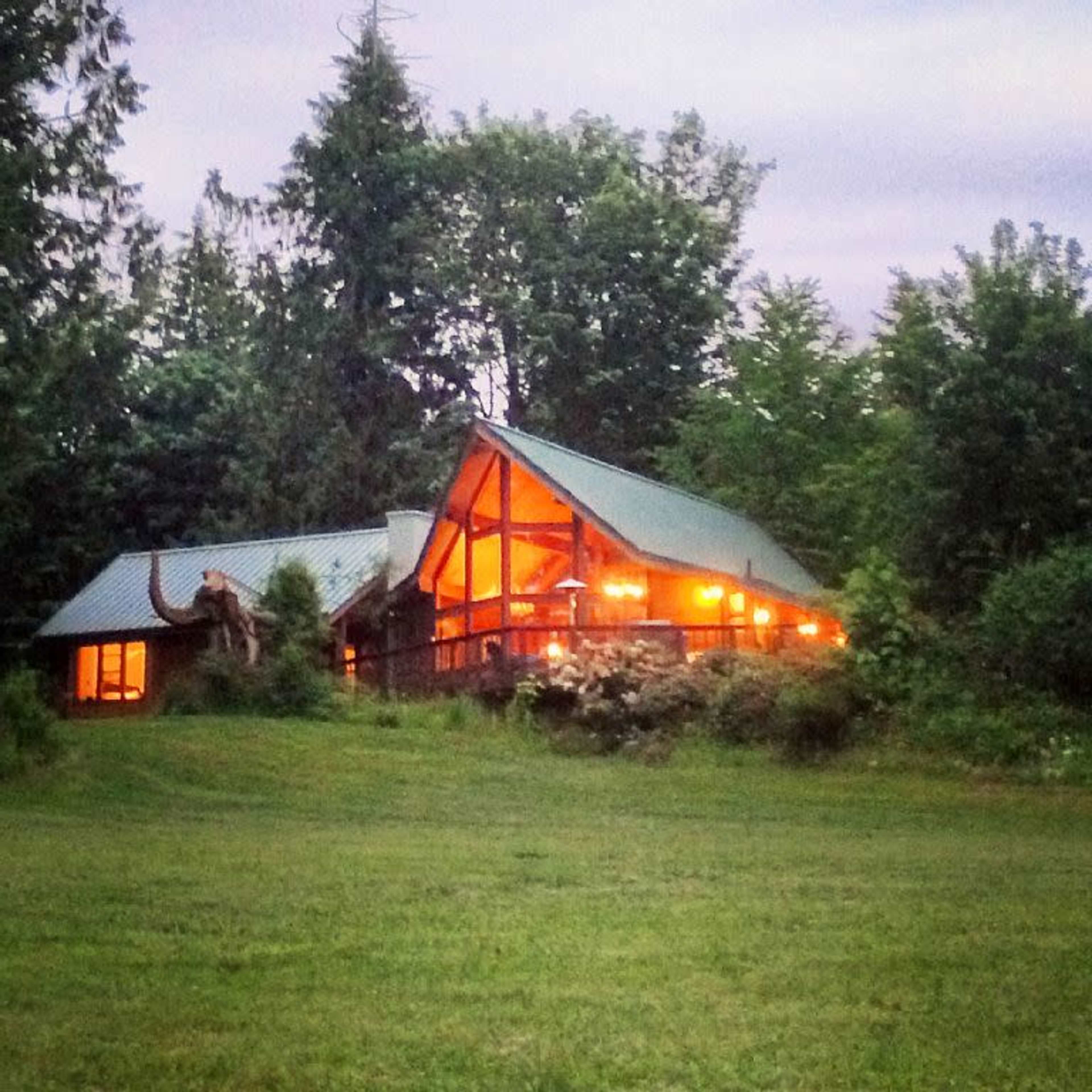 A rustic cabin with a sloped roof is illuminated by warm lights, surrounded by trees and greenery in a twilight setting.