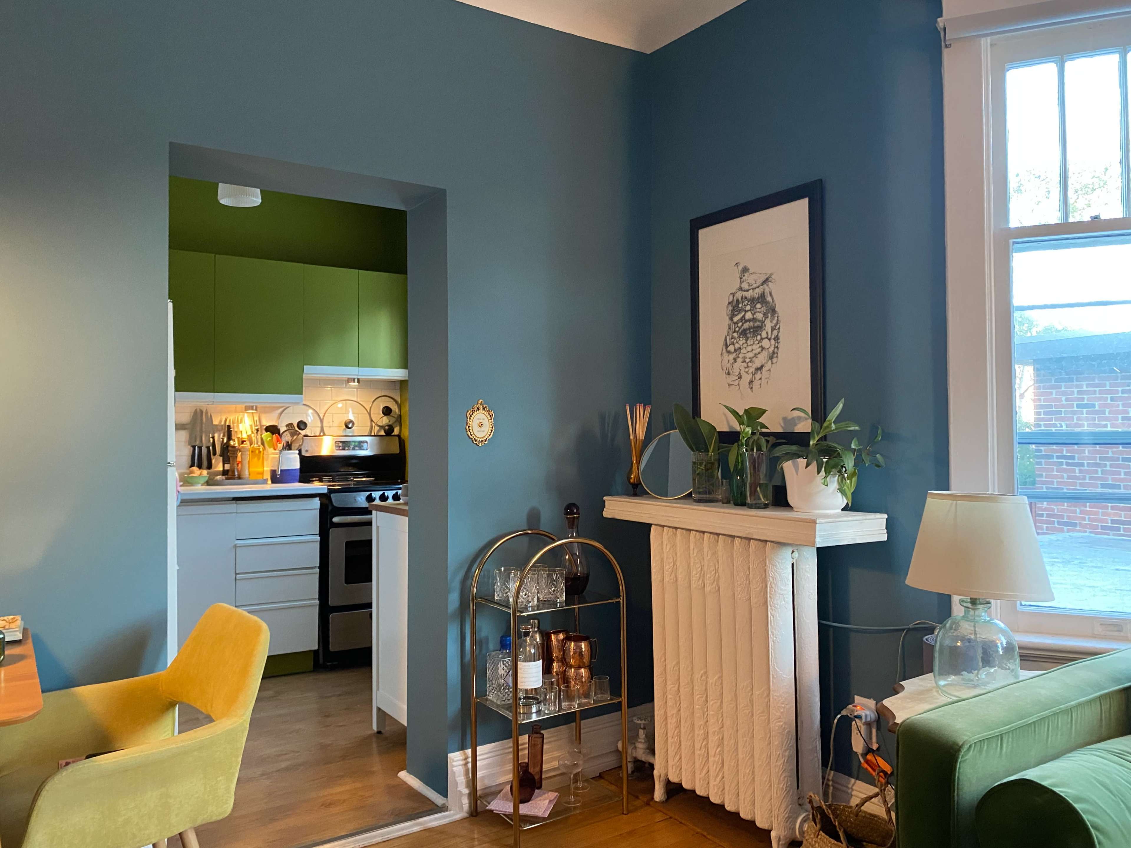 A cozy room with a kitchen area featuring green cabinets, a small bar cart with drinks, and a white radiator beside a large window.
