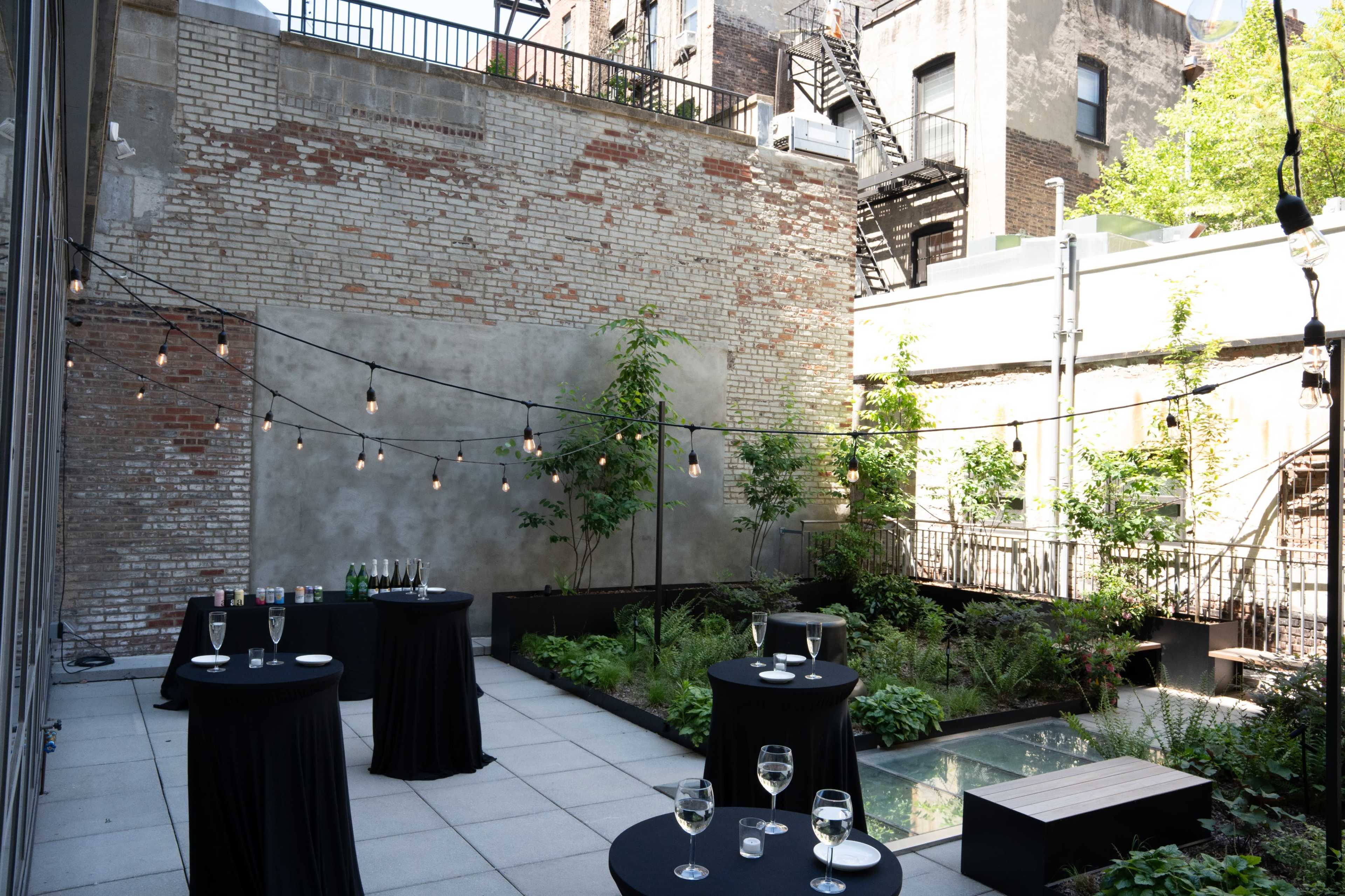The image shows a rooftop terrace with several round tables covered in black cloth, surrounded by greenery and string lights.