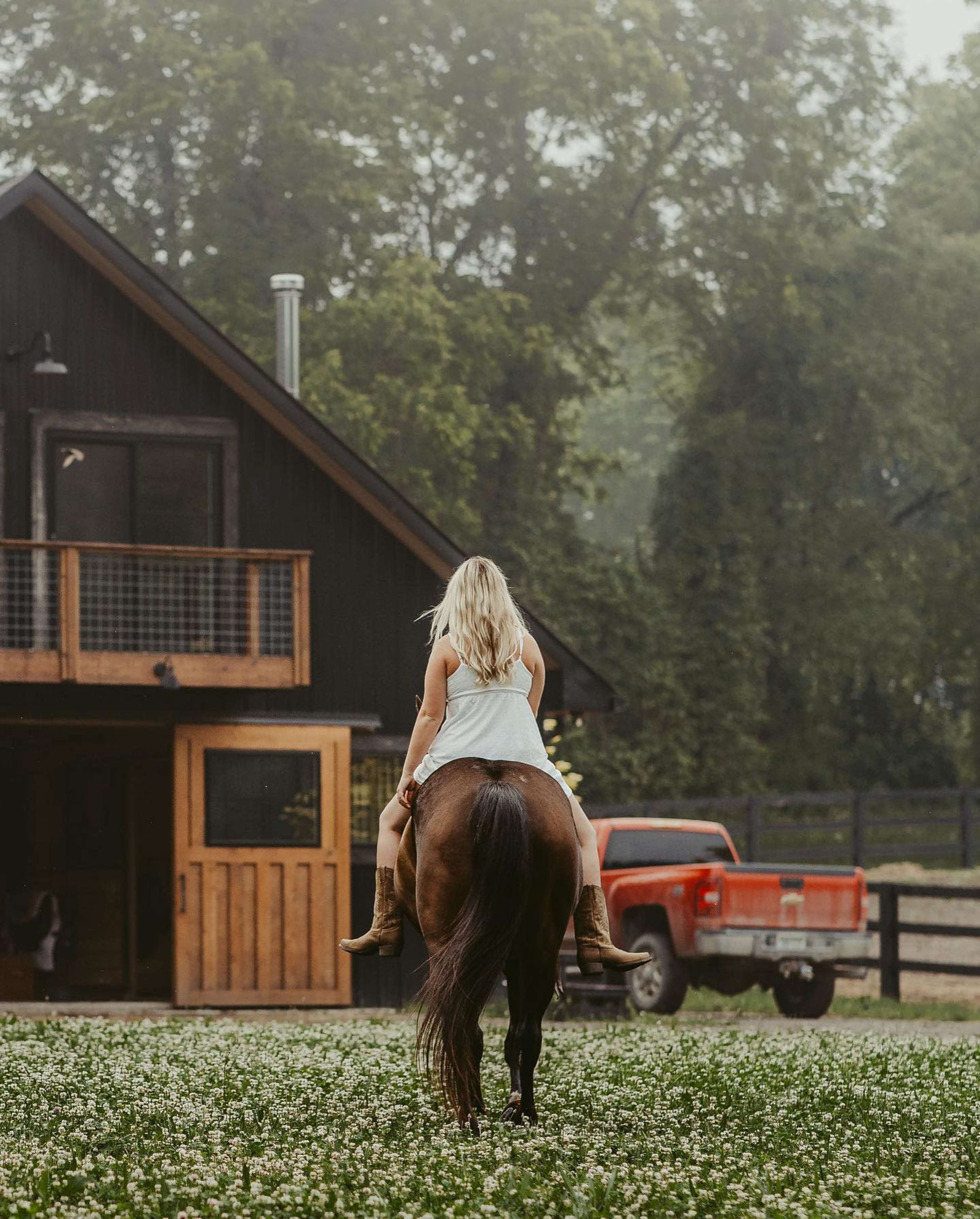 A person with long hair rides a horse in front of a wooden building and a red truck in a grassy area.