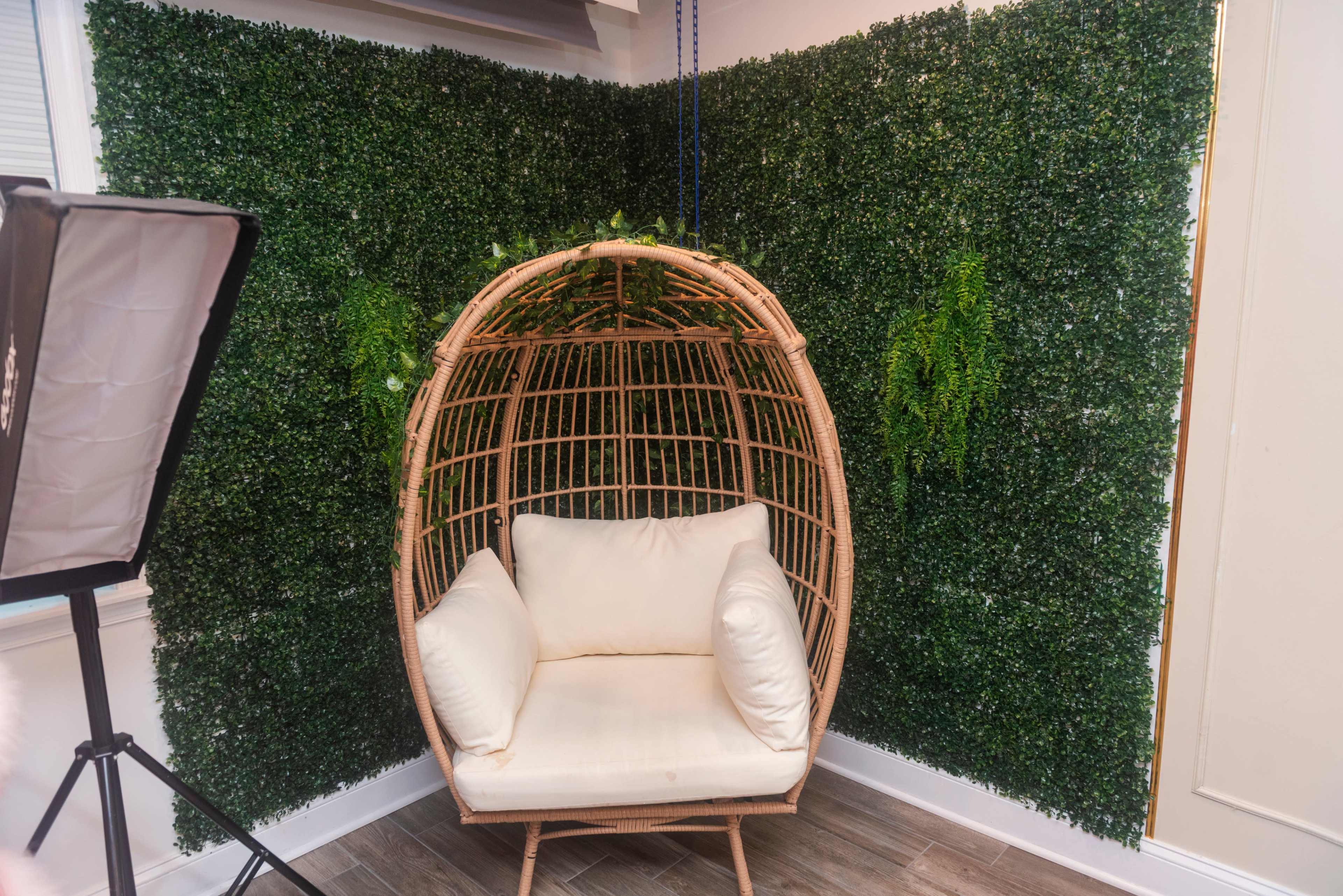 A hanging wicker chair with cushions is positioned against a wall covered in artificial greenery.