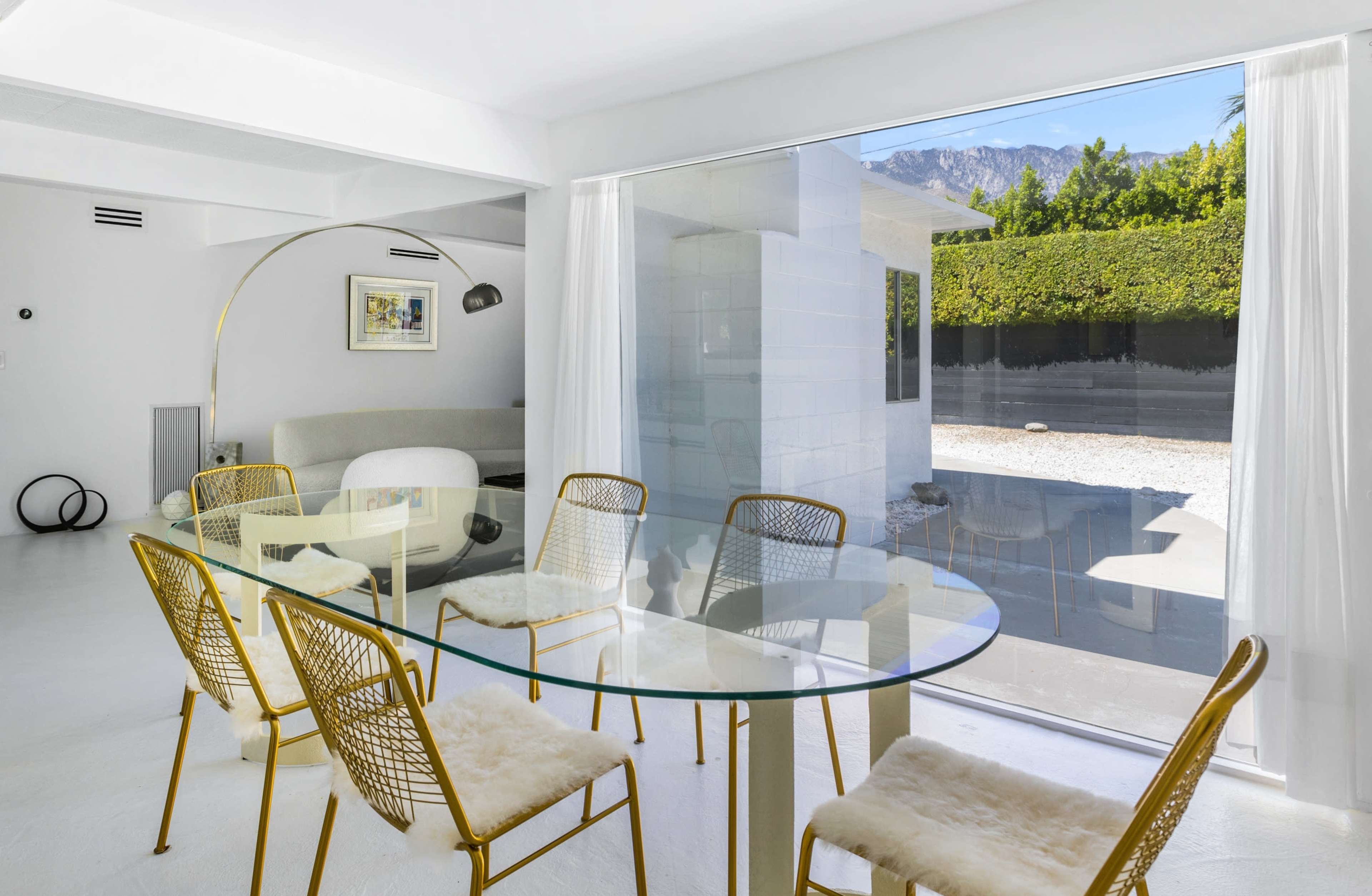 A modern dining area with a glass table surrounded by gold chairs, next to large windows overlooking a landscaped outdoor space.