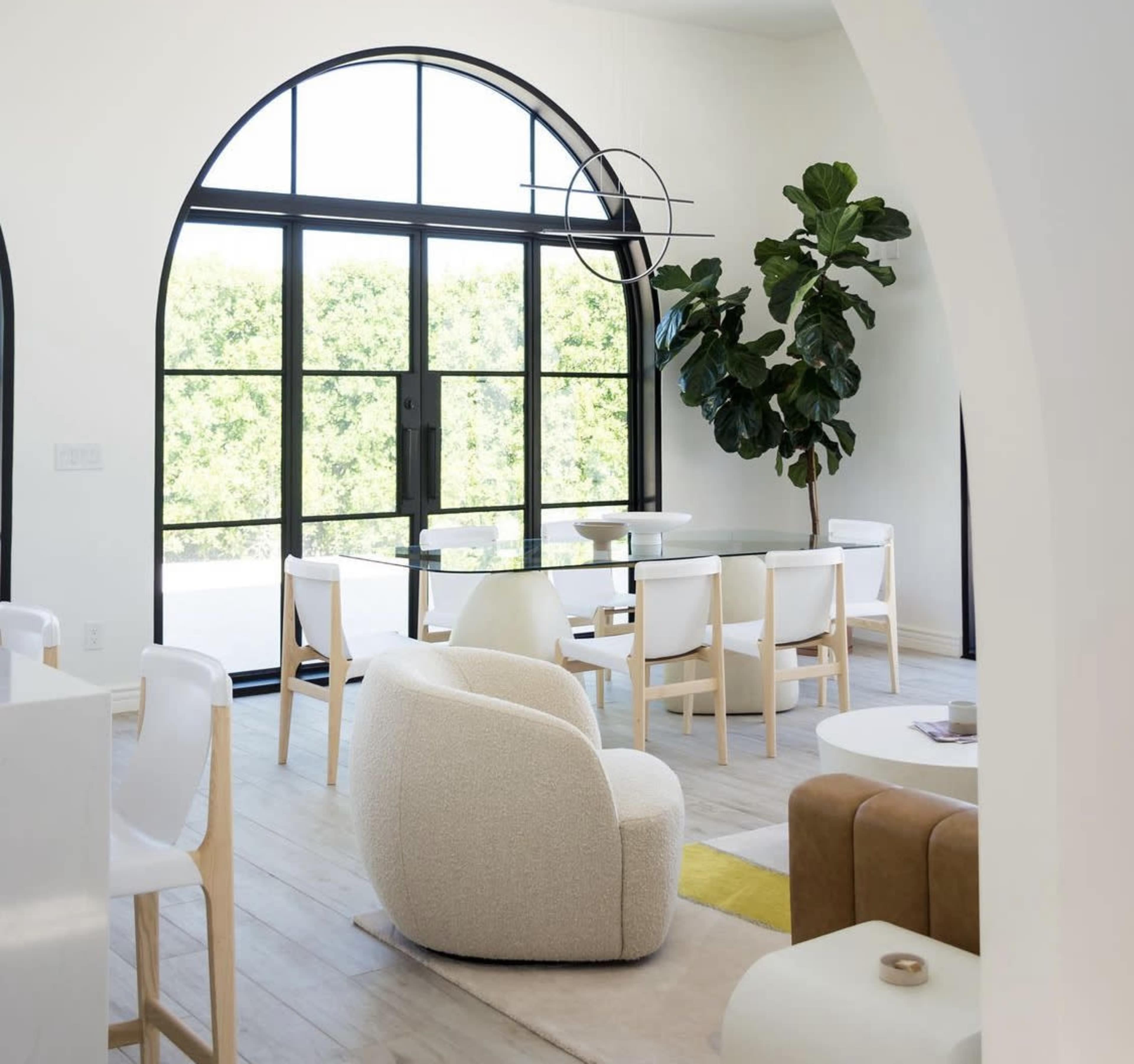 A modern interior space featuring a large arched window, a dining table with white chairs, and a potted plant in a light-filled room.