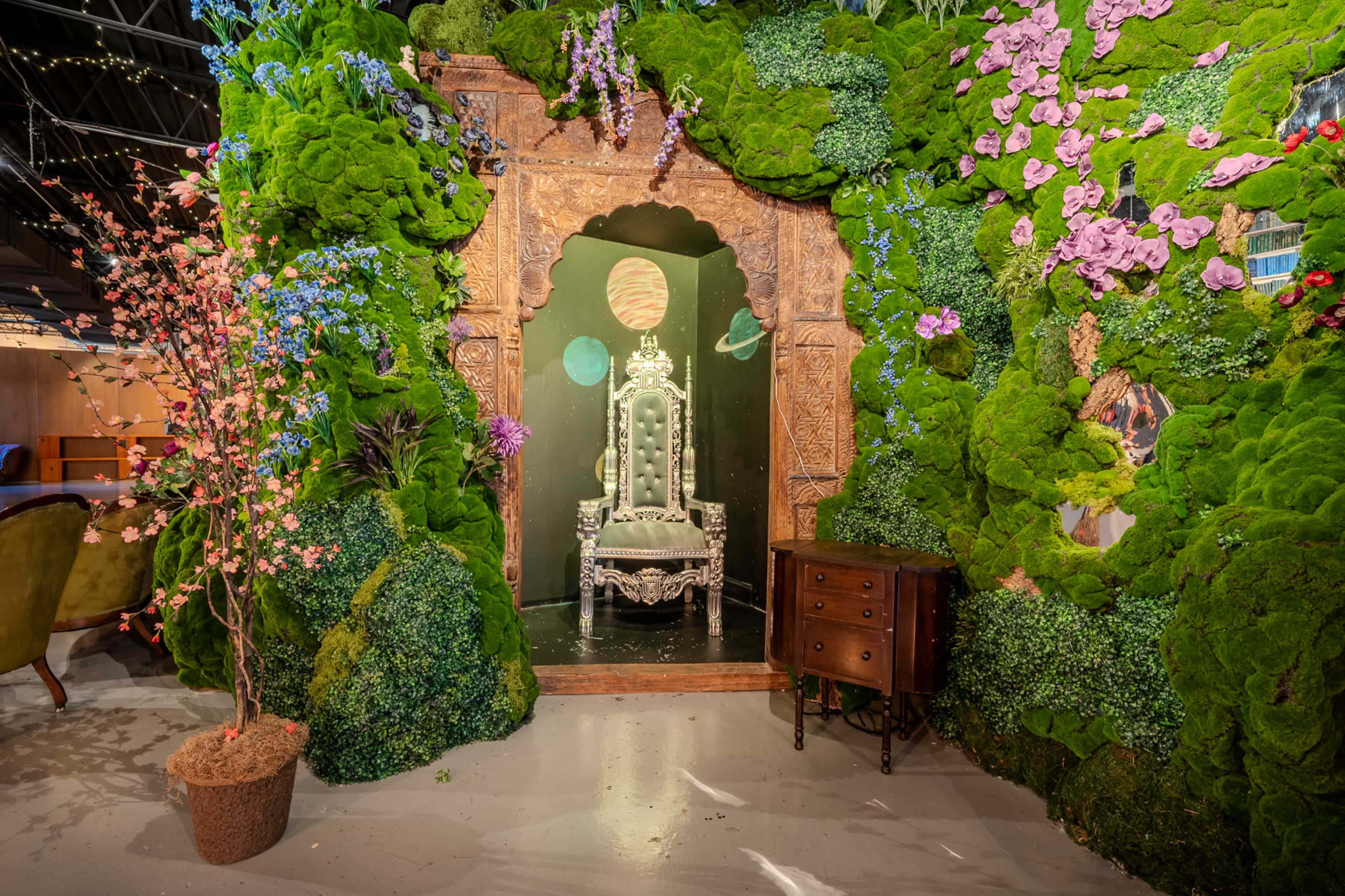 The image features an ornate throne set against a backdrop of lush greenery and colorful flowers within a decorative archway.