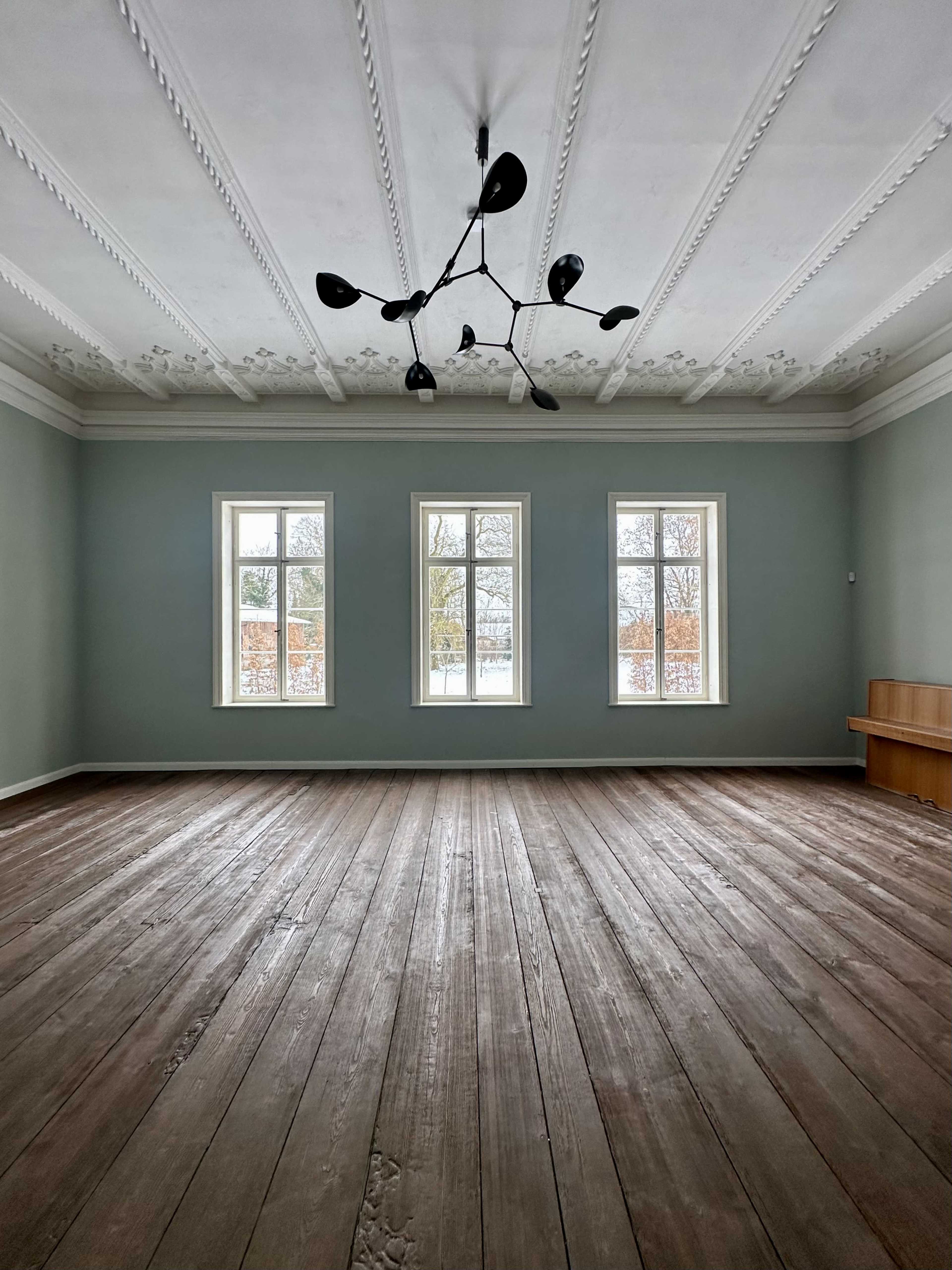 The image shows a spacious room with light green walls, wooden flooring, three windows, and a modern chandelier.