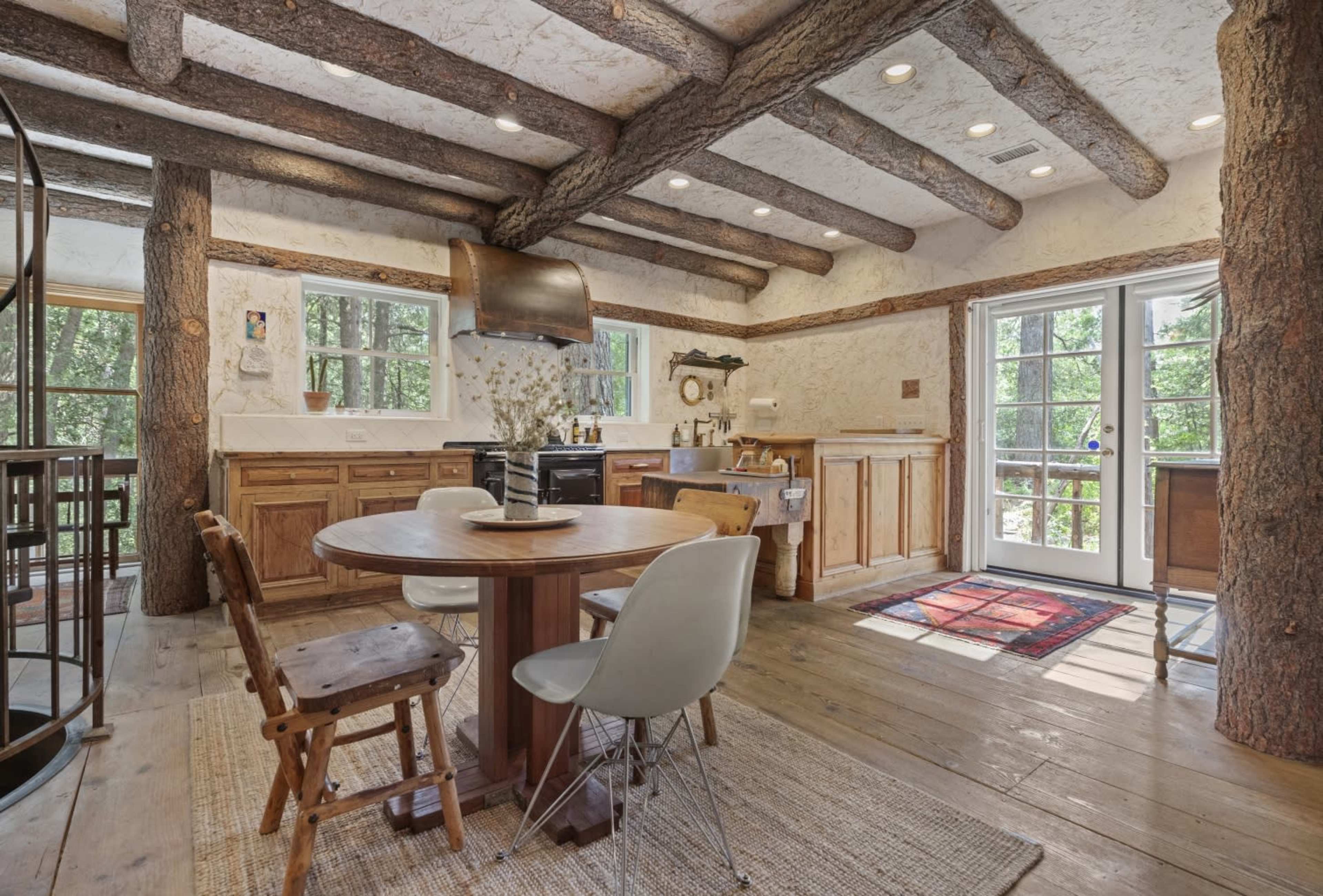 The image depicts a rustic kitchen with wooden beams, a round dining table, and large windows that open to a view of the outdoors.