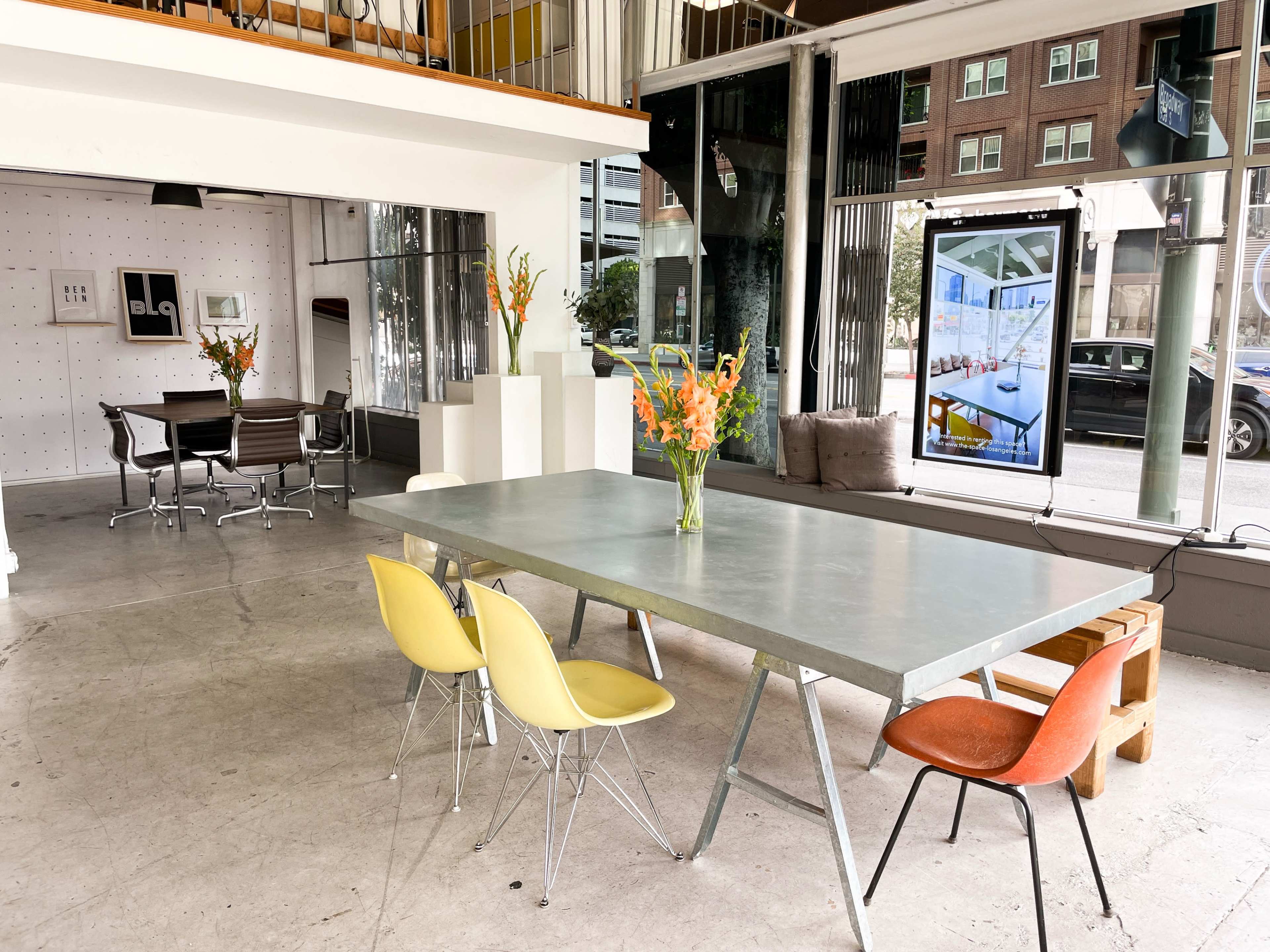 A spacious modern interior features a large metal dining table surrounded by colorful chairs, with a glass partition leading to a separate meeting area.
