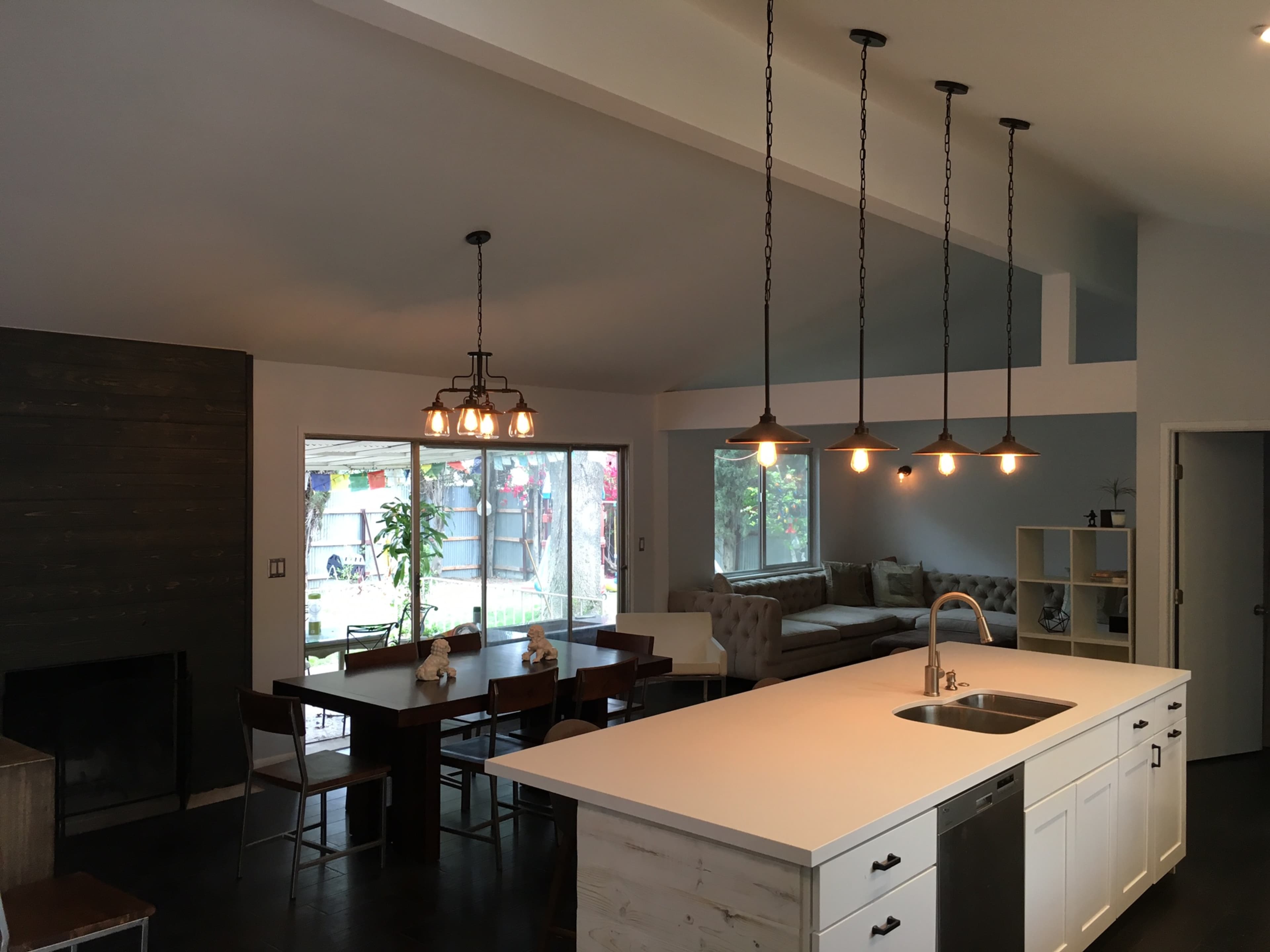 The image shows an open-concept kitchen and living area with a large island, wooden dining table, and a sectional sofa, illuminated by several hanging light fixtures.
