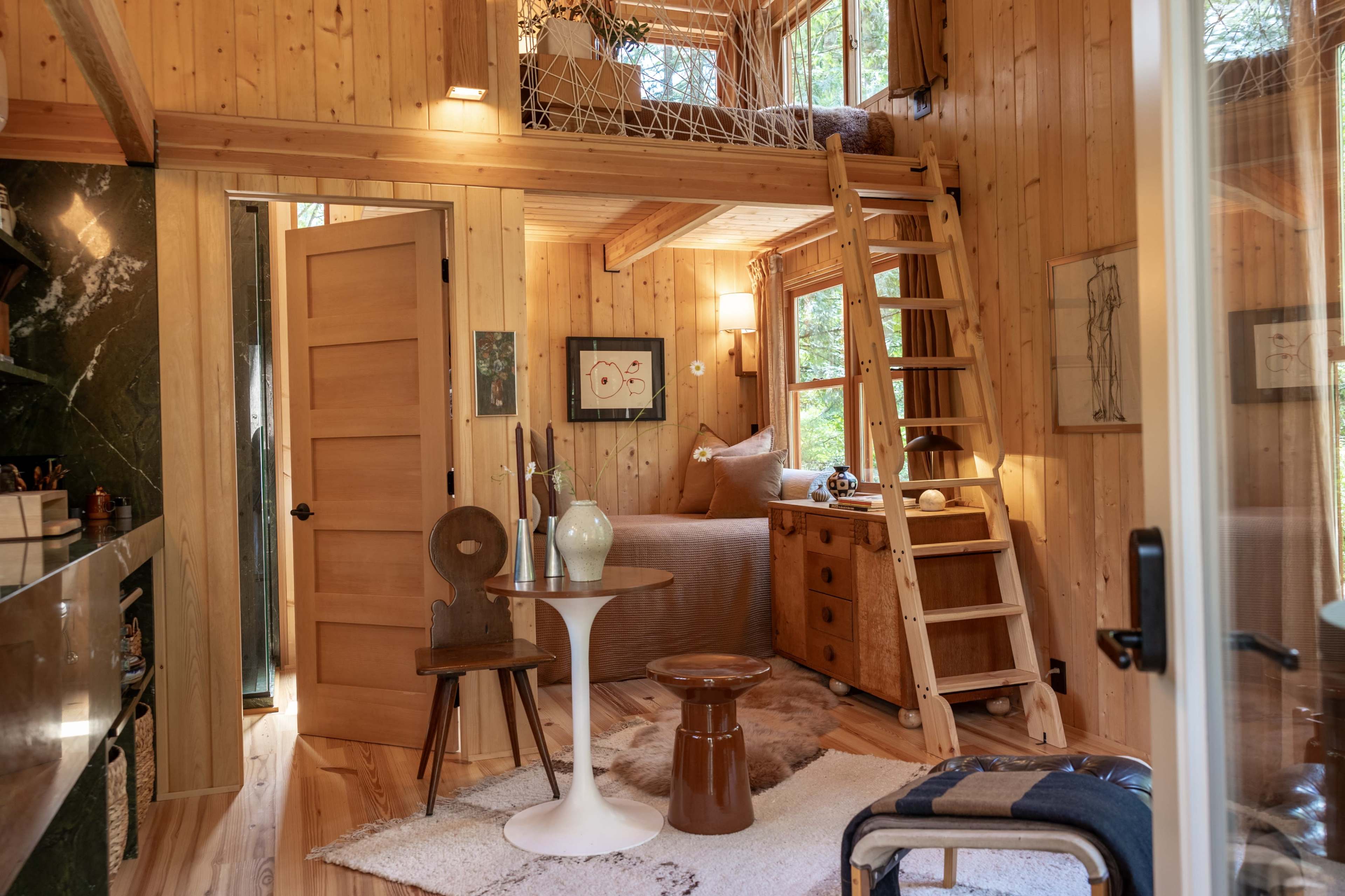 The image shows a wooden interior of a small cabin, featuring a loft accessible by a ladder, a seating area with a table and chairs, and decorative plants.