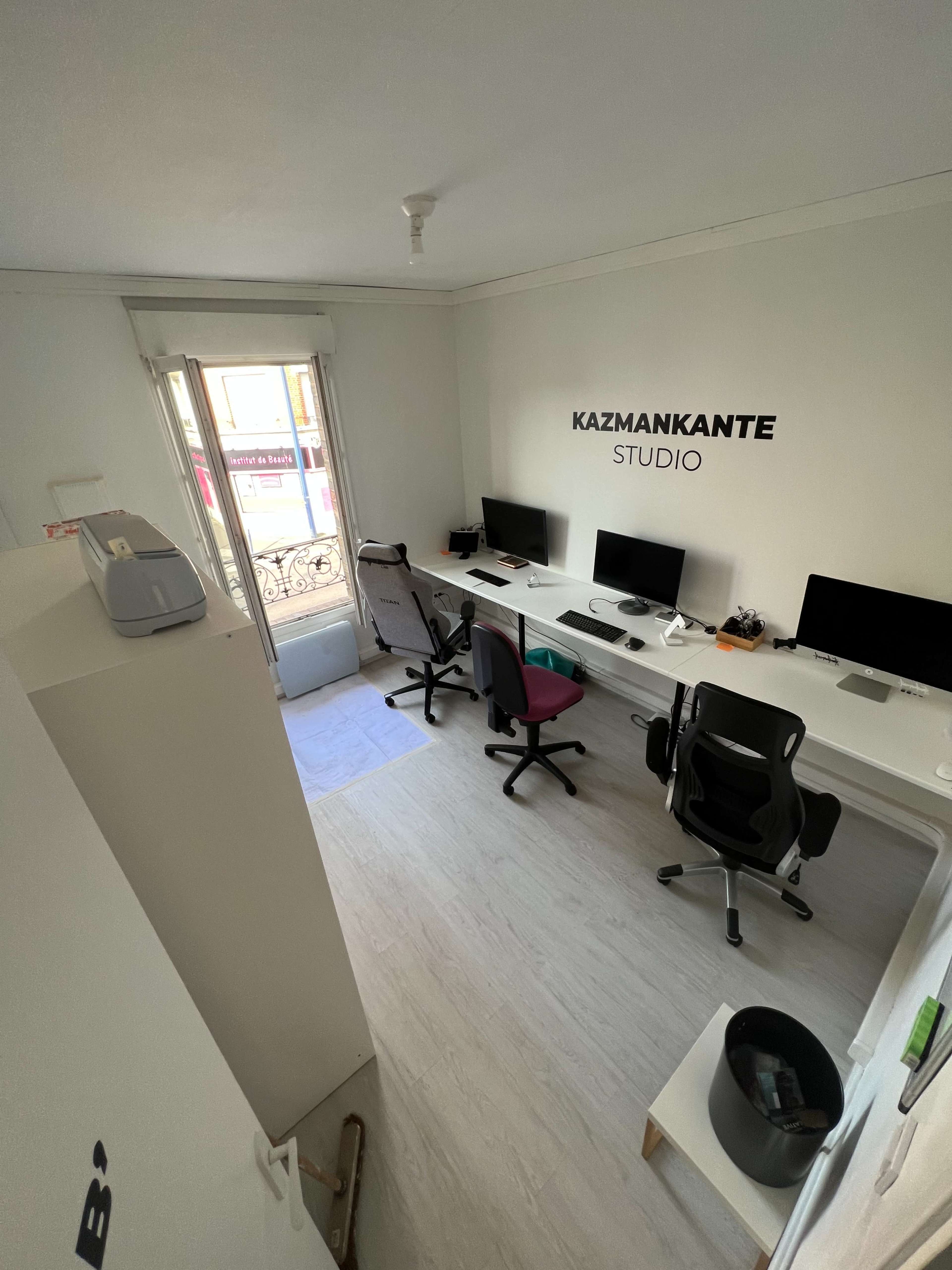 A minimalist office space with several desks, computers, and a window overlooking a balcony, featuring the name "KAZMANKANTE STUDIO" on the wall.