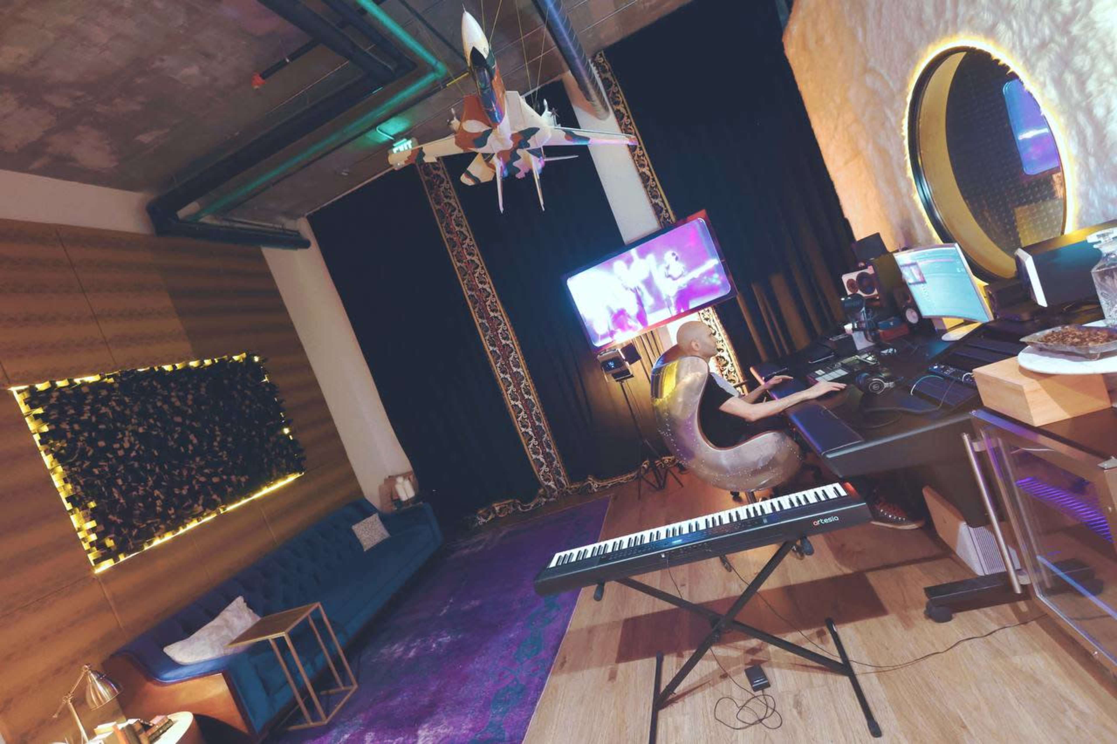 A person is sitting at a workstation with a keyboard and multiple screens in a modern studio featuring a sofa, decorative wall, and large monitor.