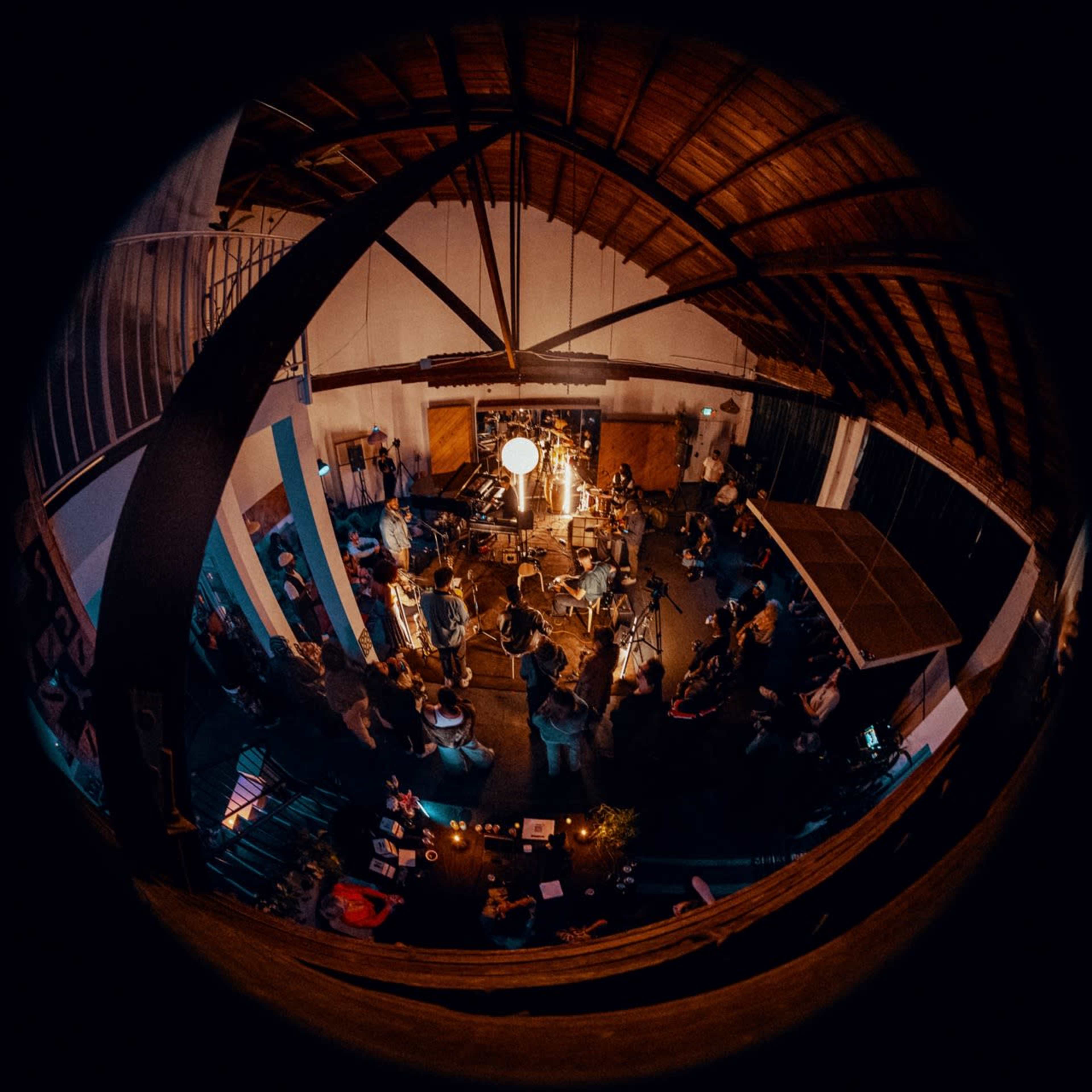 A group of people is gathered in a well-lit, spacious interior for a performance or recording session, with various instruments and a large light source at the center.
