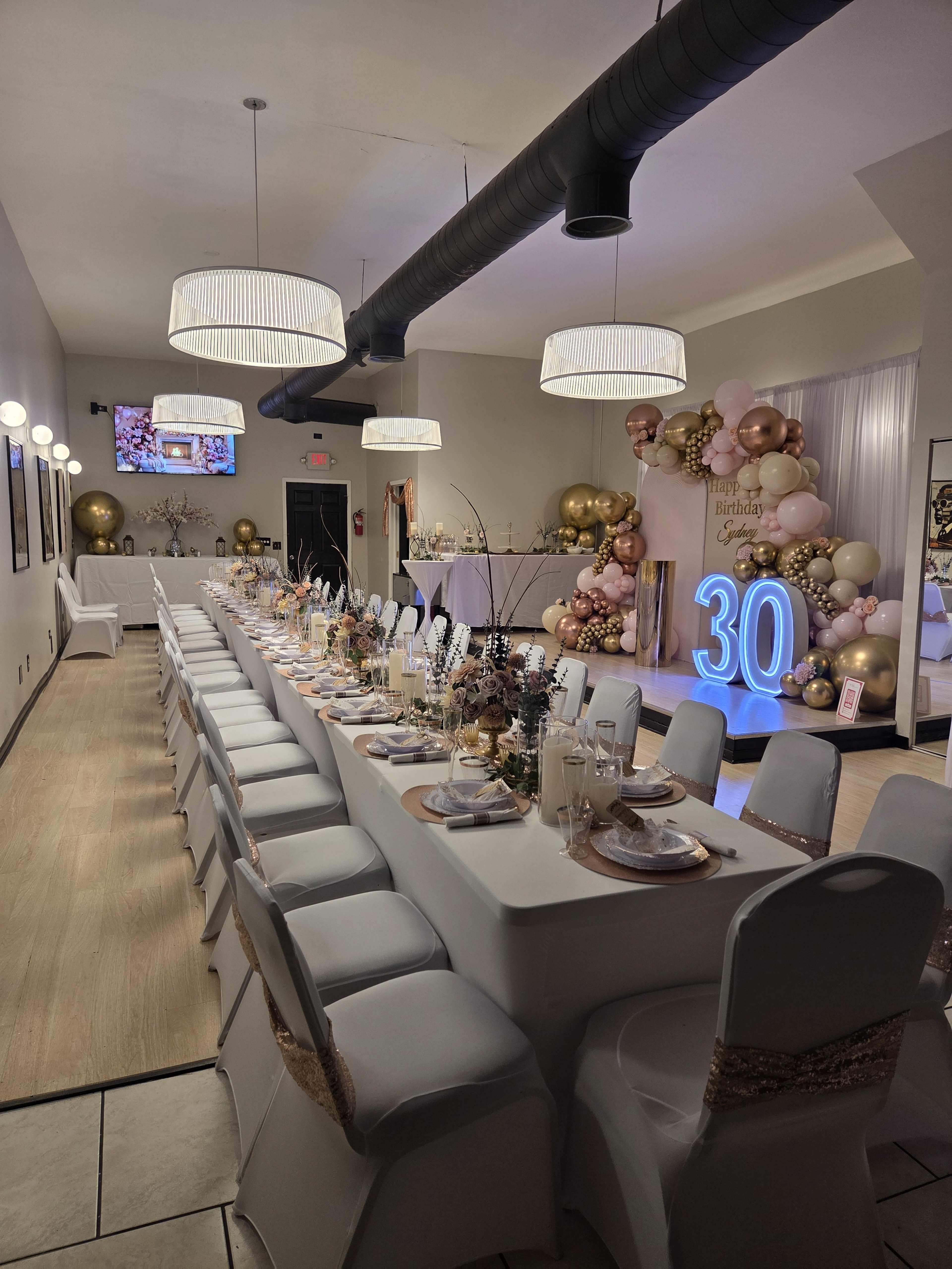 A long, elegantly set dining table is arranged in a decorated event space for a 30th birthday celebration, featuring balloons, gold accents, and a lit number "30" backdrop.