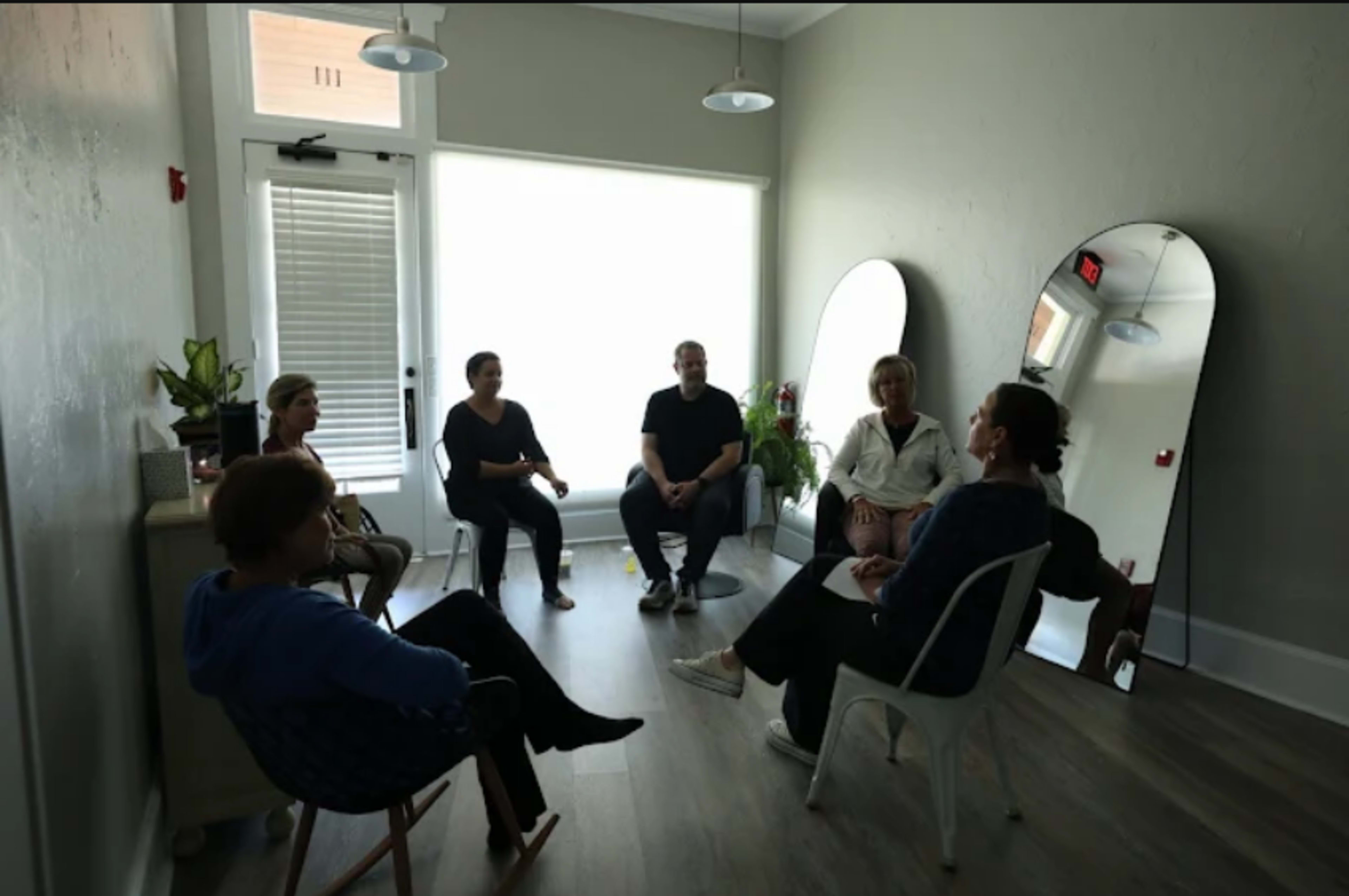 A group of six people sits in a circular arrangement within a well-lit room featuring large mirrors and indoor plants.