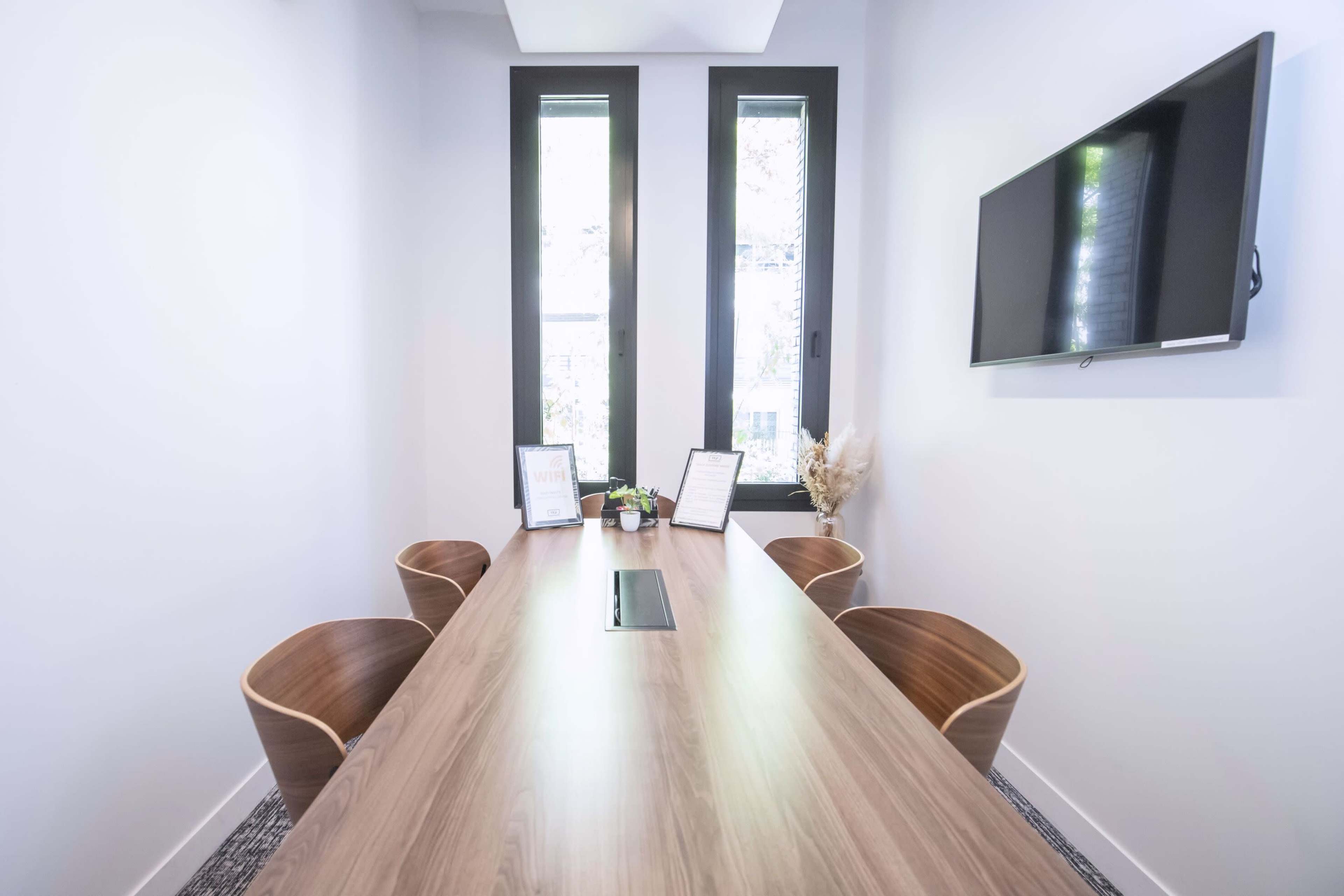 A conference room features a long wooden table surrounded by four chairs, with two large windows and a wall-mounted television.
