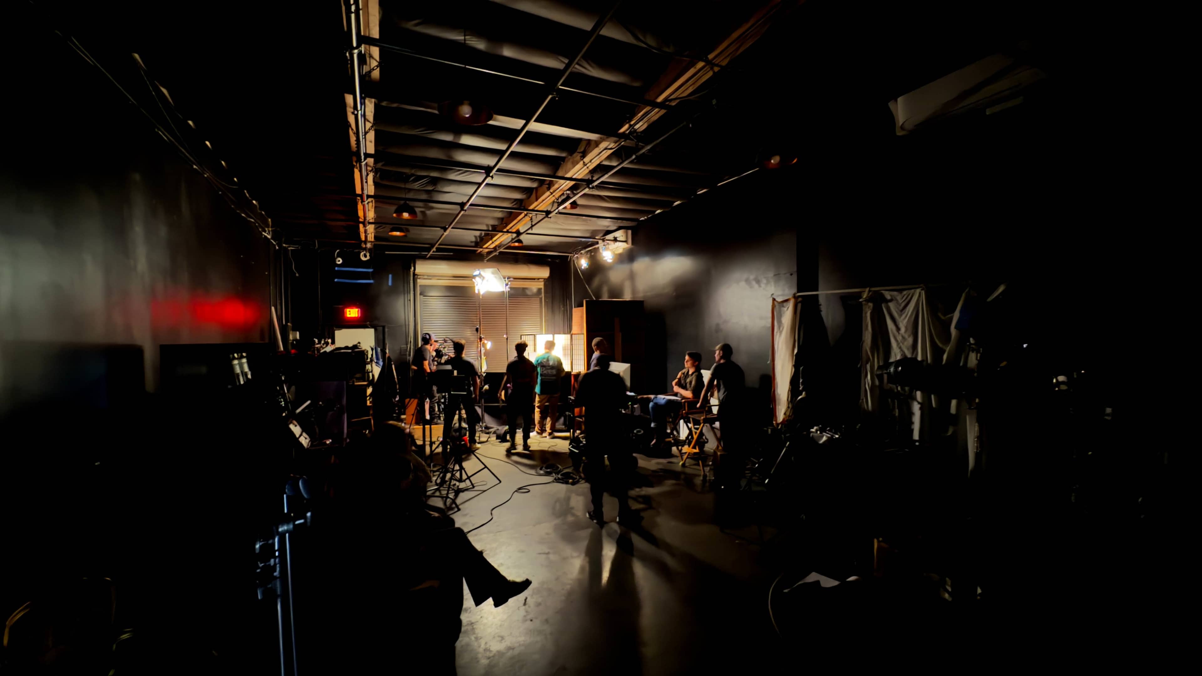 A film crew works on a set in a dimly lit studio, surrounded by equipment and various tools.