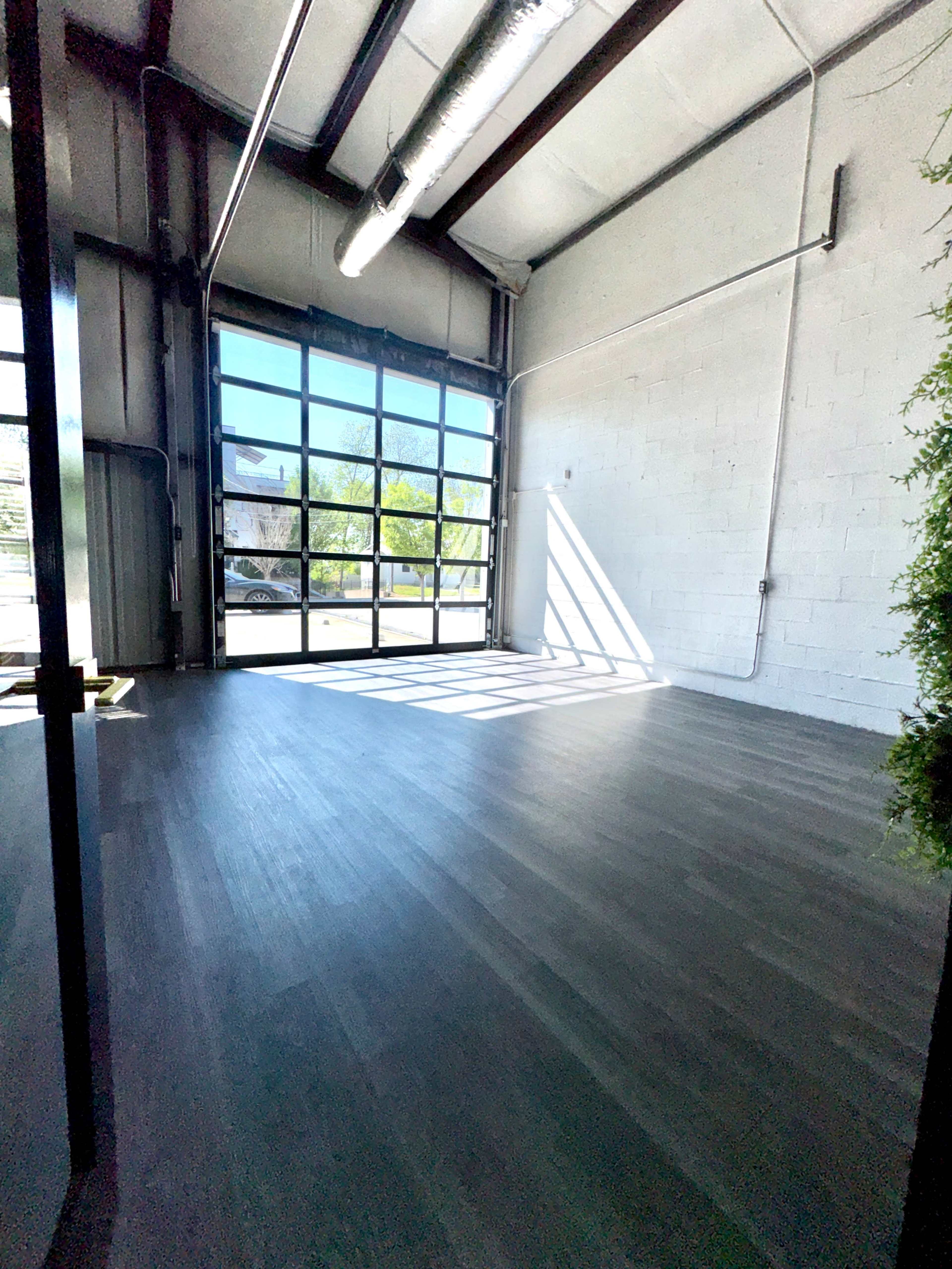 The image shows an empty room with a large glass garage door, allowing natural light to stream in, and featuring wooden floors and white walls.