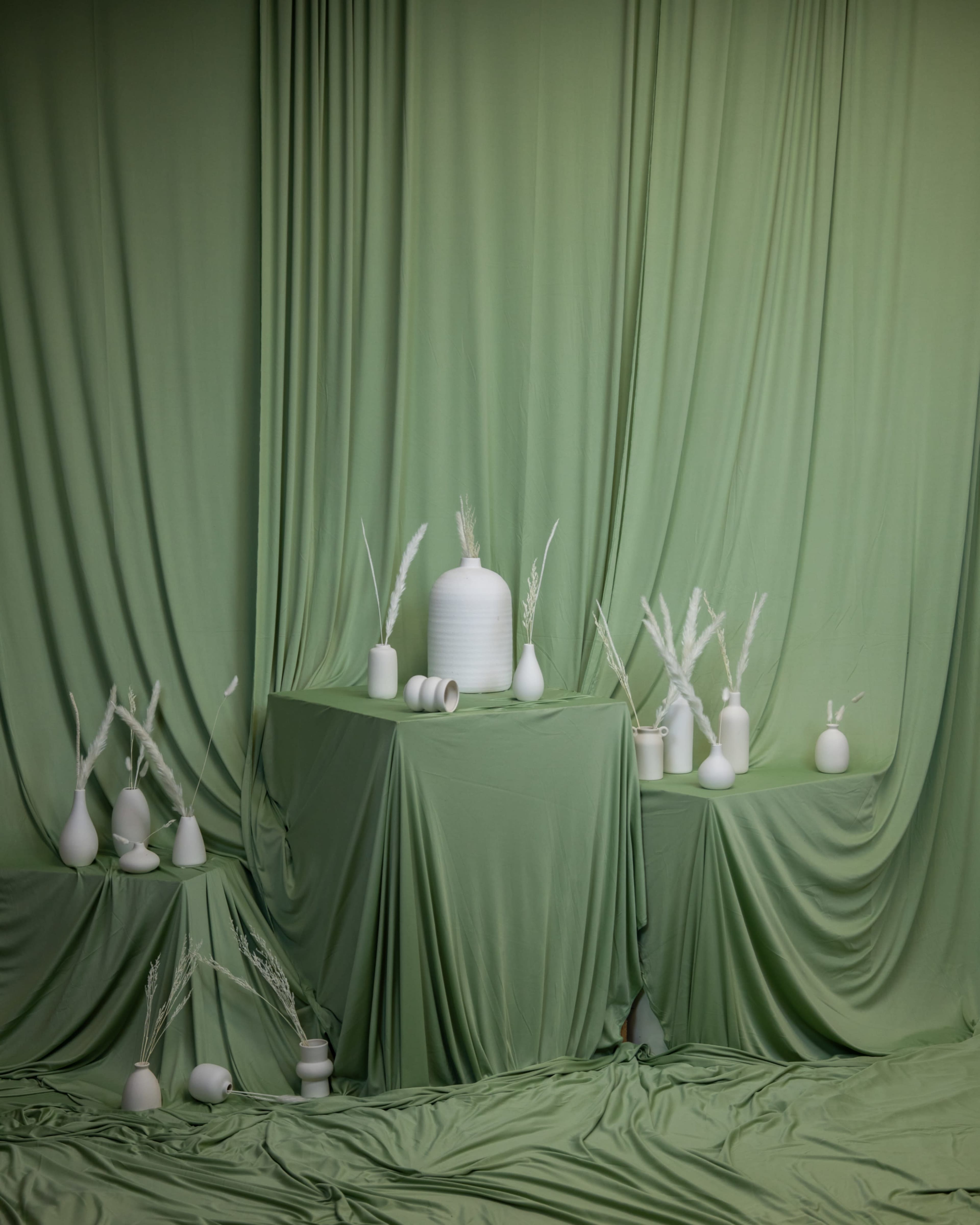 The image features a styled display with white vases and decorative elements arranged on two green-draped tables.