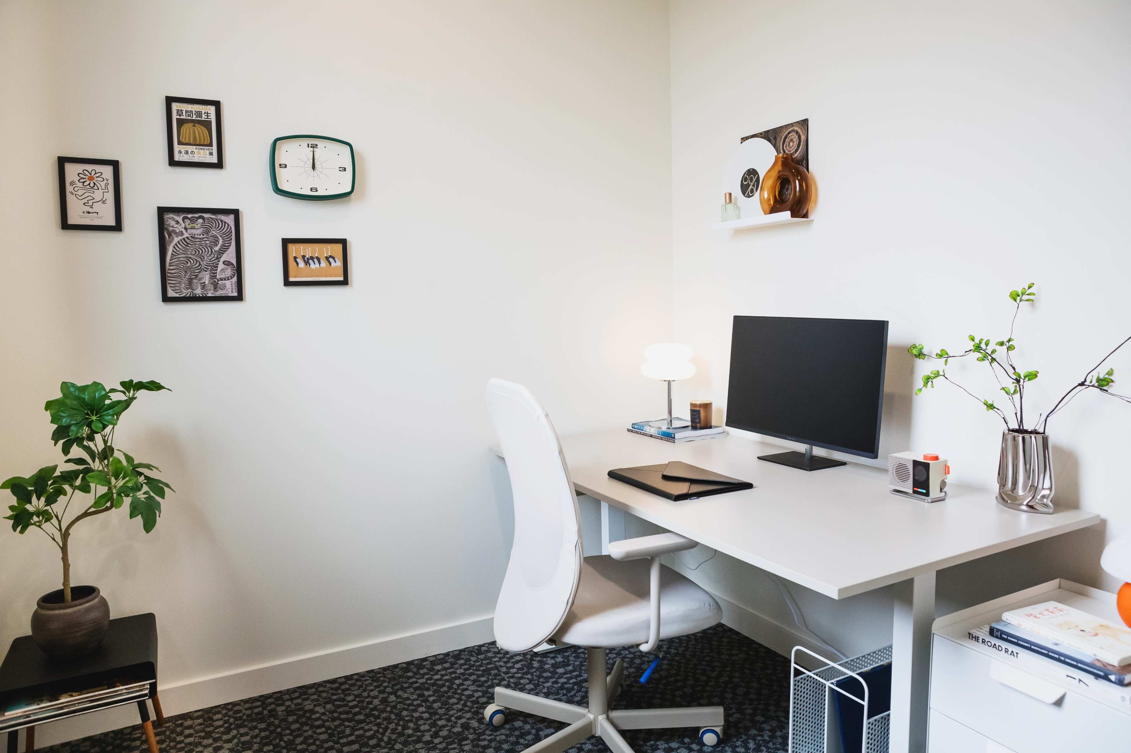 A modern office space features a desk with a computer, a white ergonomic chair, and wall art alongside a potted plant and decorative items on a shelf.