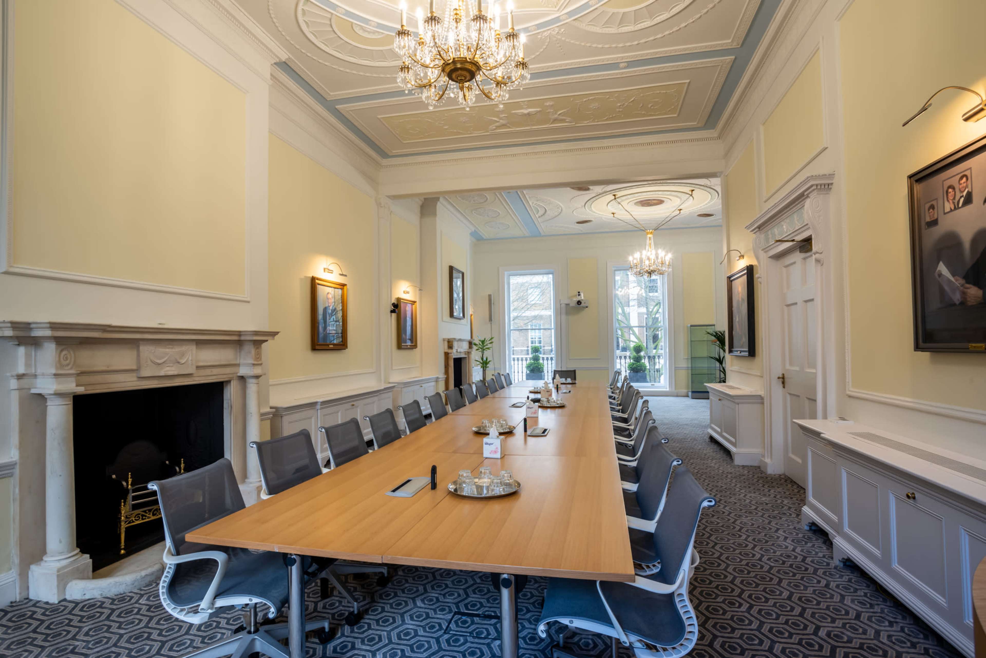 A spacious conference room features a long wooden table surrounded by modern chairs, with large windows allowing natural light and ornate chandeliers overhead.