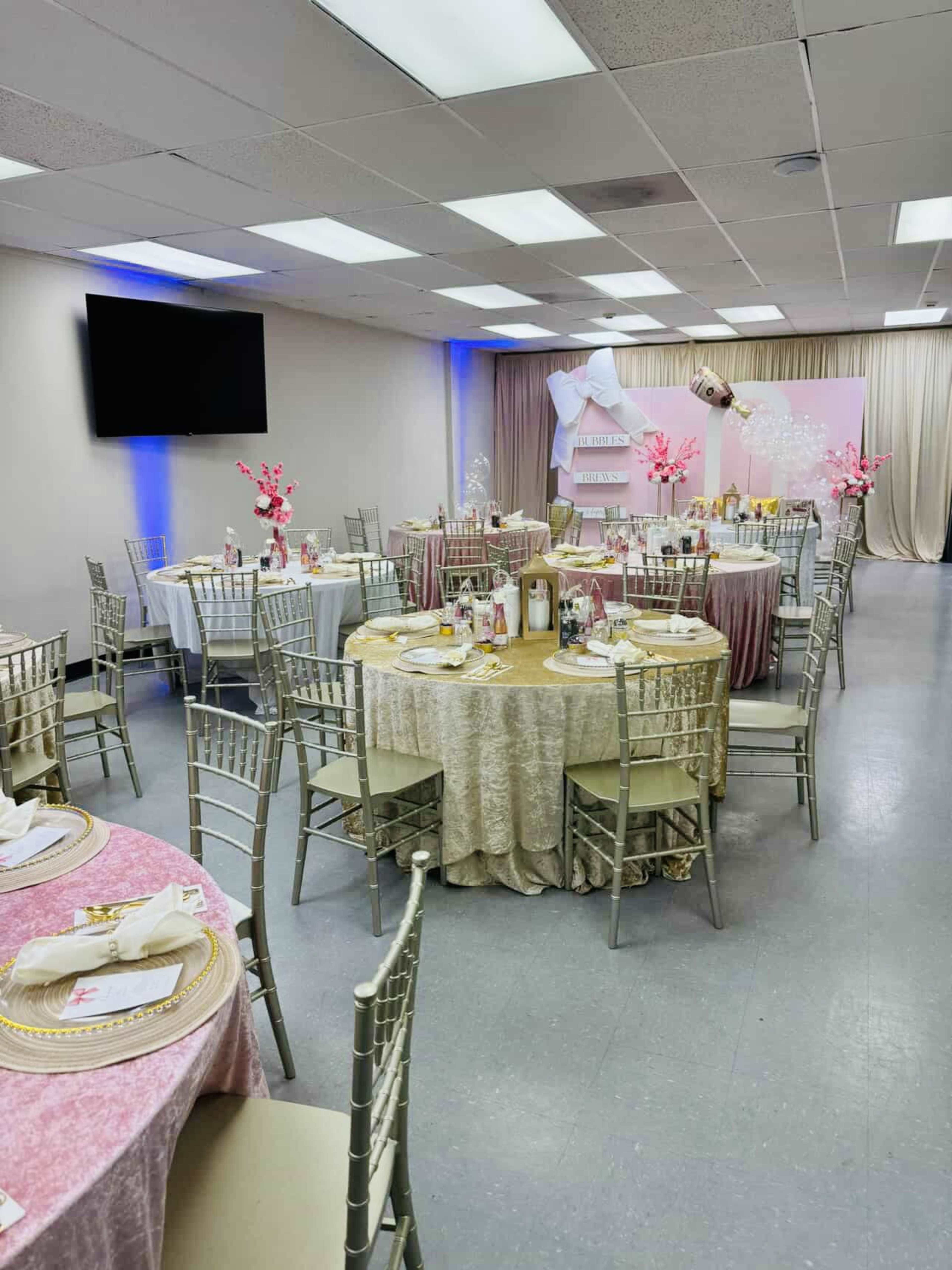 A decorated banquet hall features several circular tables with tablecloths in various colors, each set with plates and centerpieces, while a backdrop displays elegant decor.