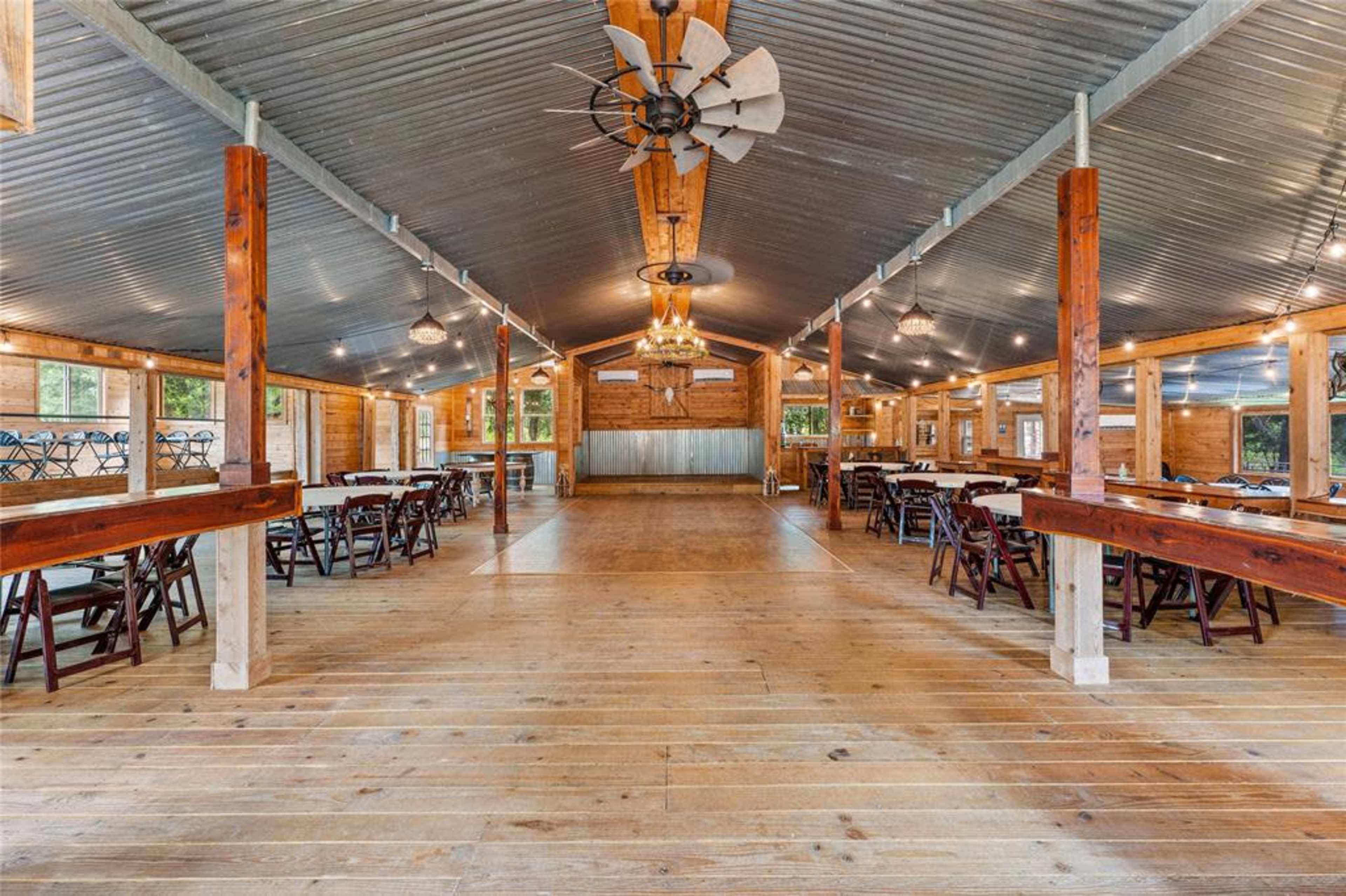 A large, open event space with wooden floors, metal roofing, and tables arranged along the sides.
