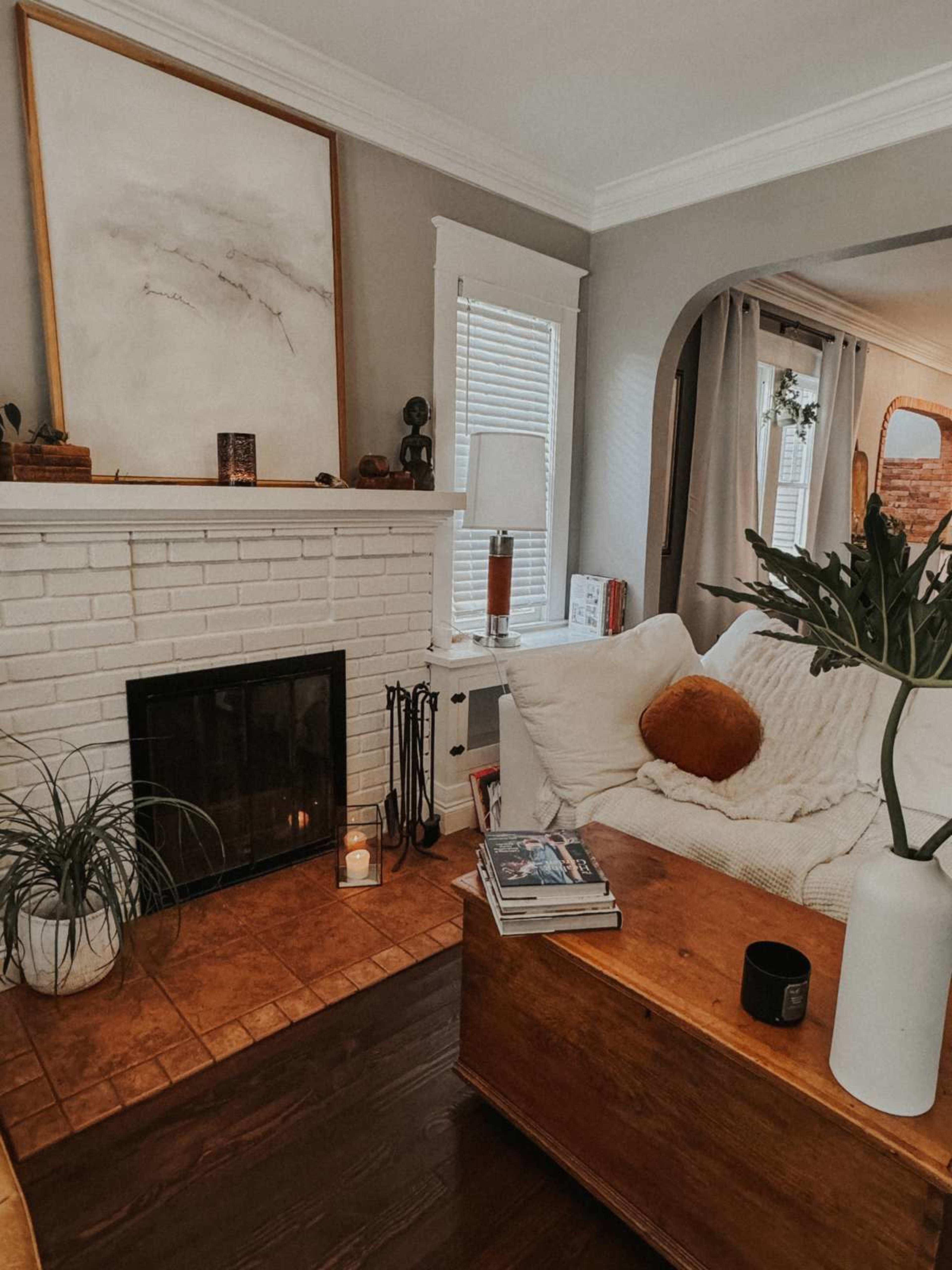 The living room features a white-brick fireplace, a cozy sofa with an orange pillow, a wooden coffee table, and decorative elements arranged around the space.