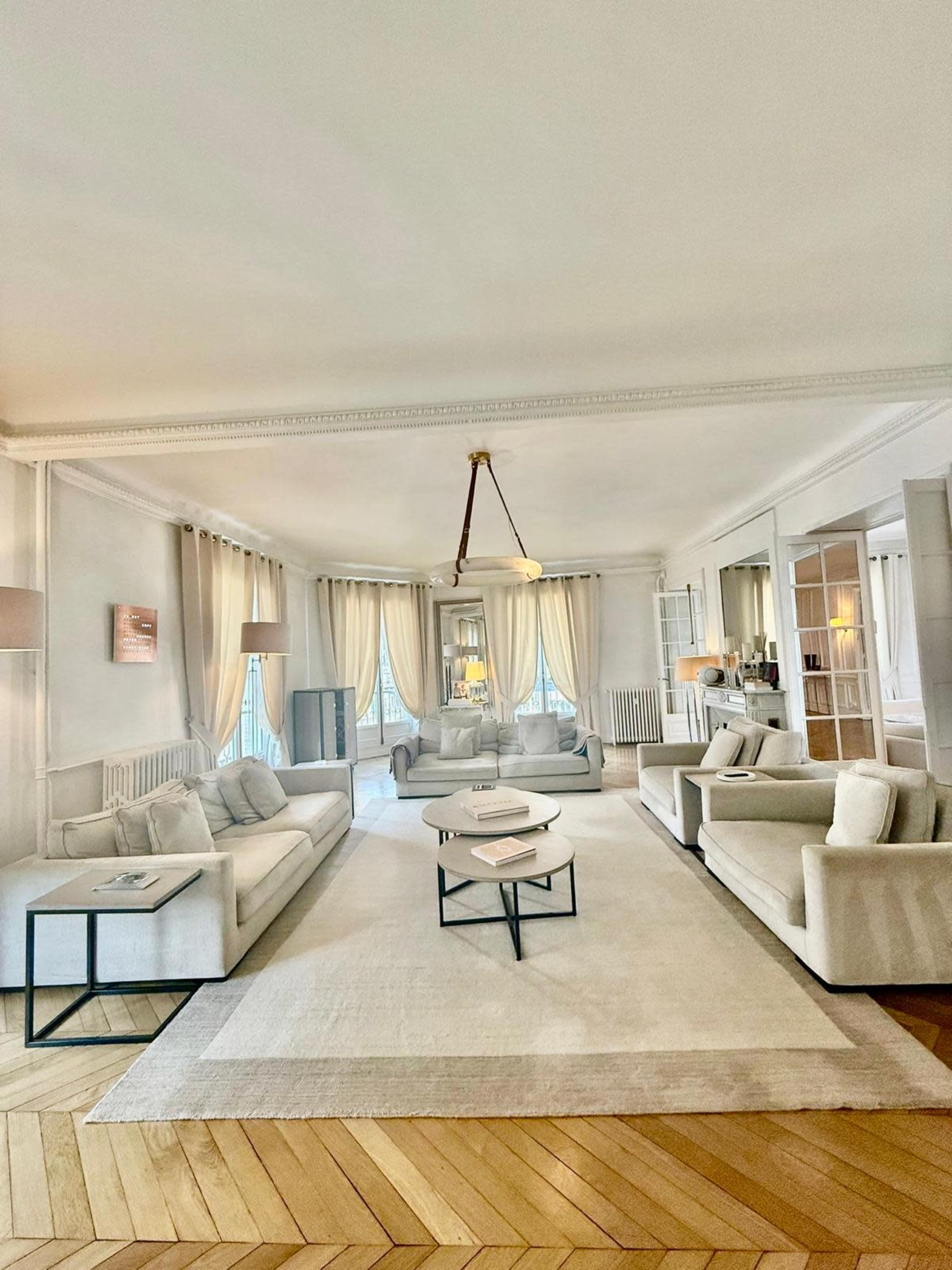 The image shows a spacious living room with two light-colored sofas, a round coffee table, and large windows dressed with sheer curtains.