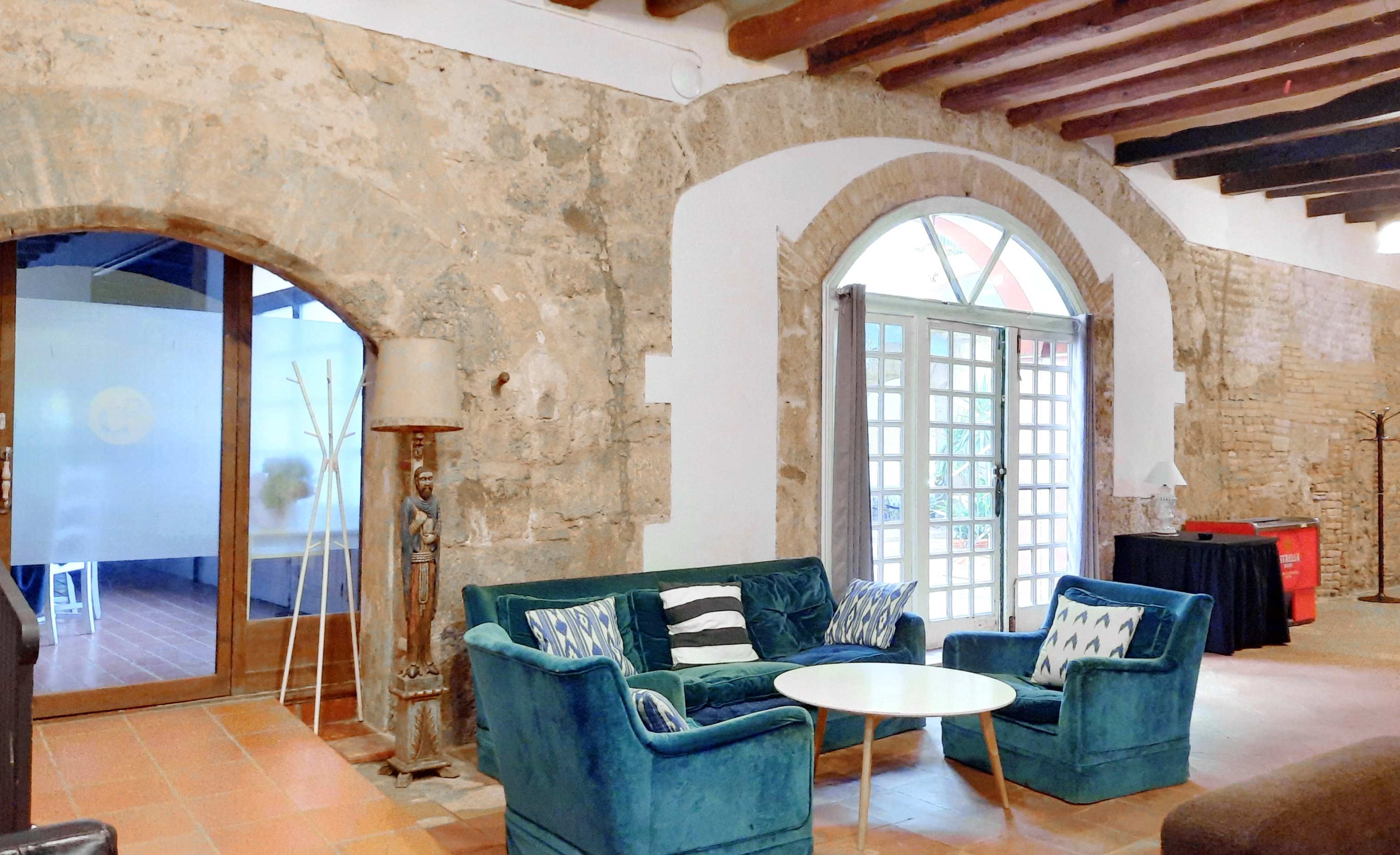 The image shows a cozy sitting area featuring a teal sofa with patterned pillows, a round coffee table, and an archway leading to a bright room with large windows.