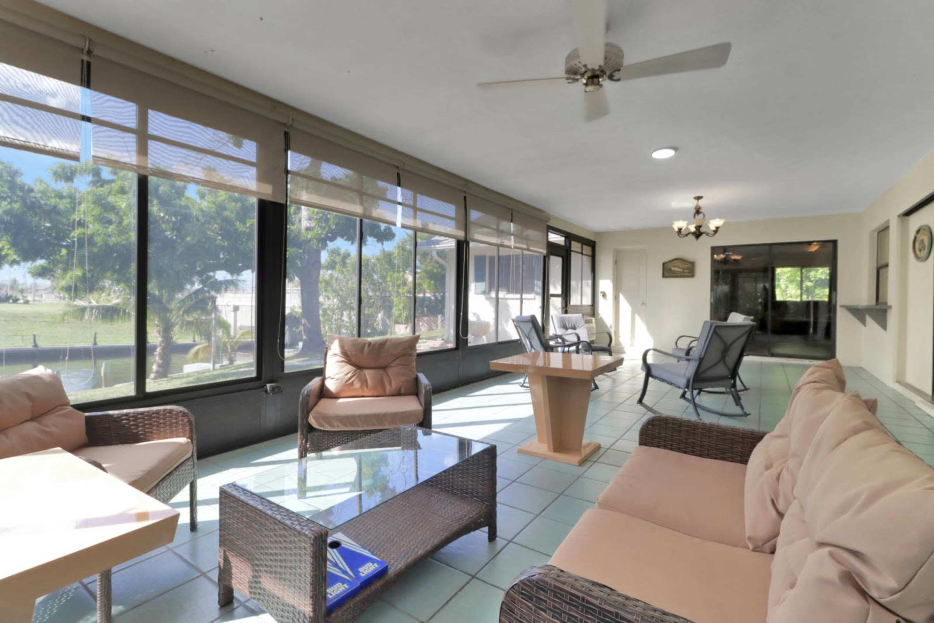The image shows a sunroom furnished with a glass-top table, comfortable seating, and large windows allowing natural light to enter.