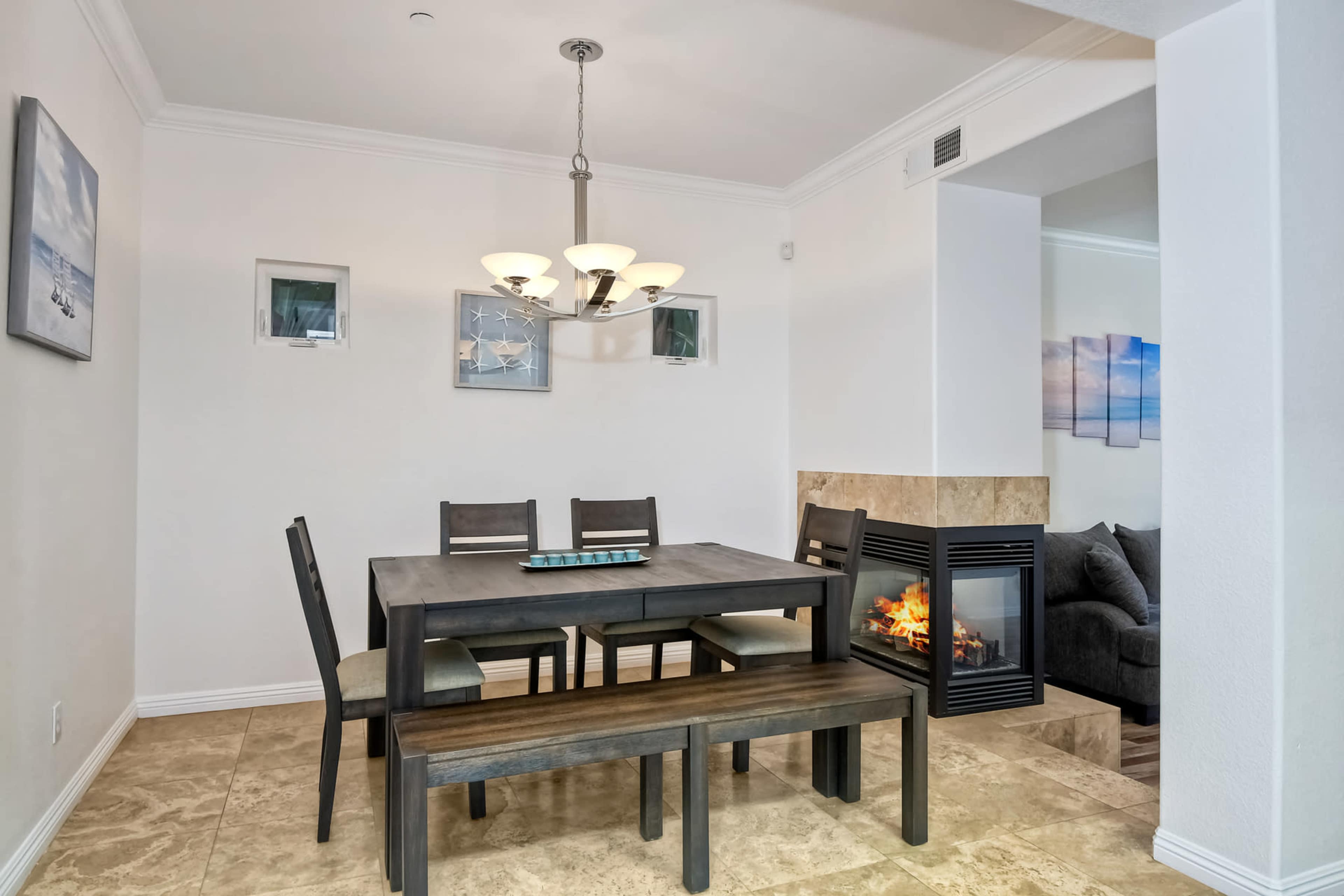 A dining area features a rectangular table with four chairs, a bench, a chandelier, and a fireplace, with artwork displayed on the walls.