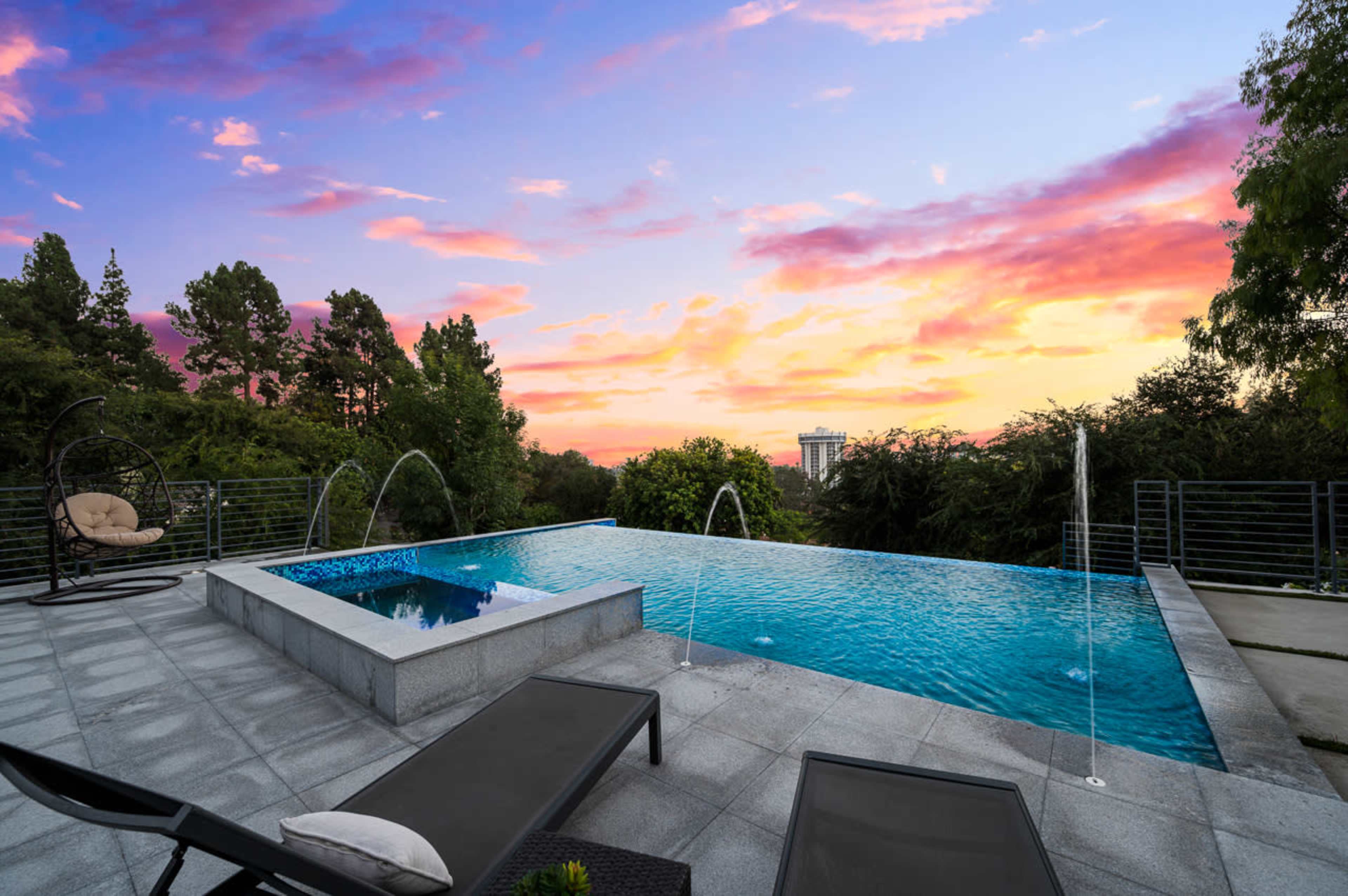 A modern swimming pool with an infinity edge, surrounded by trees and a sunset sky.